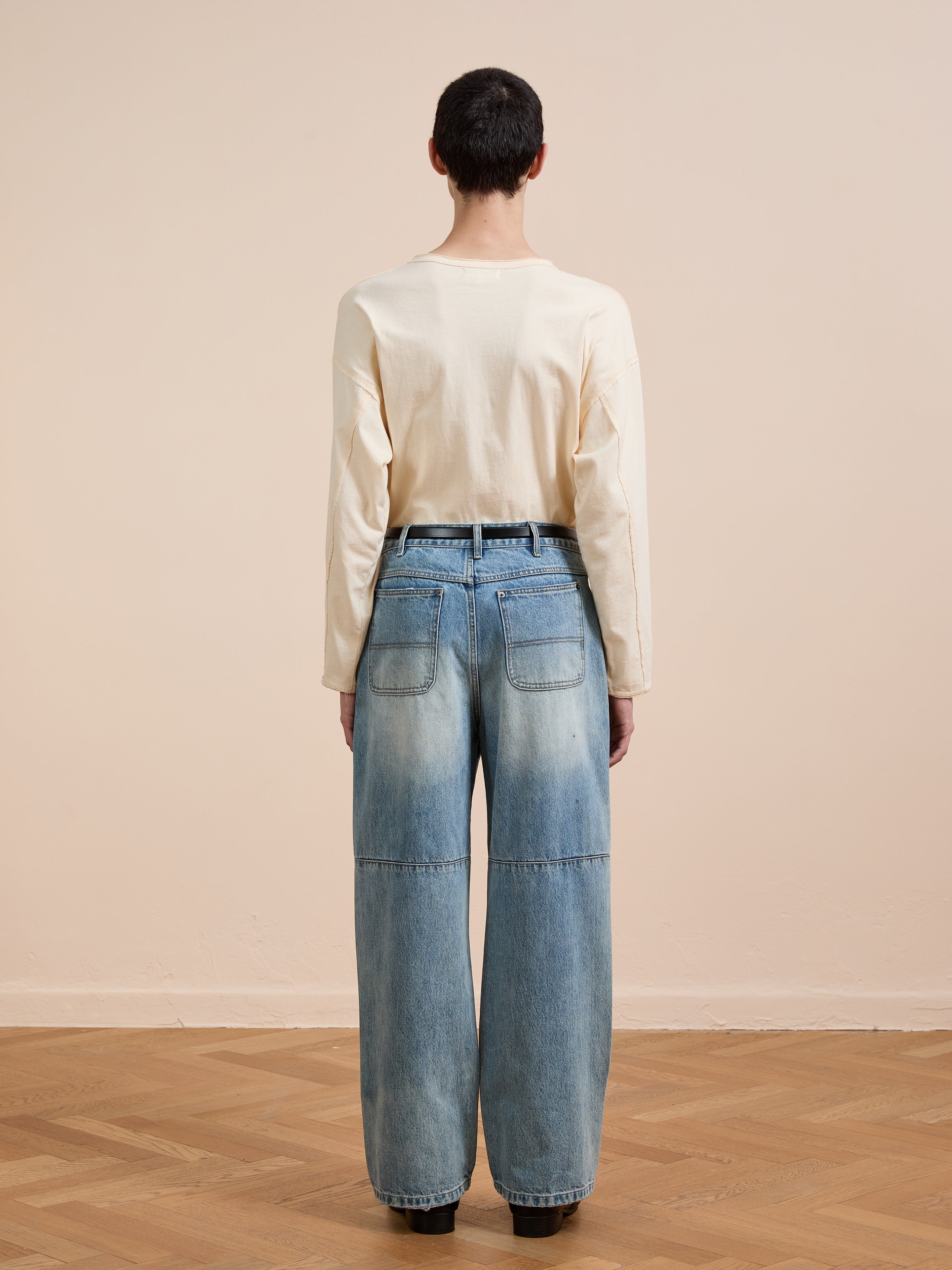 A person stands indoors, back to camera, wearing FOUND’s Hand Art Jeans and a long-sleeve cream shirt on a wooden floor with a plain beige wall in the background.