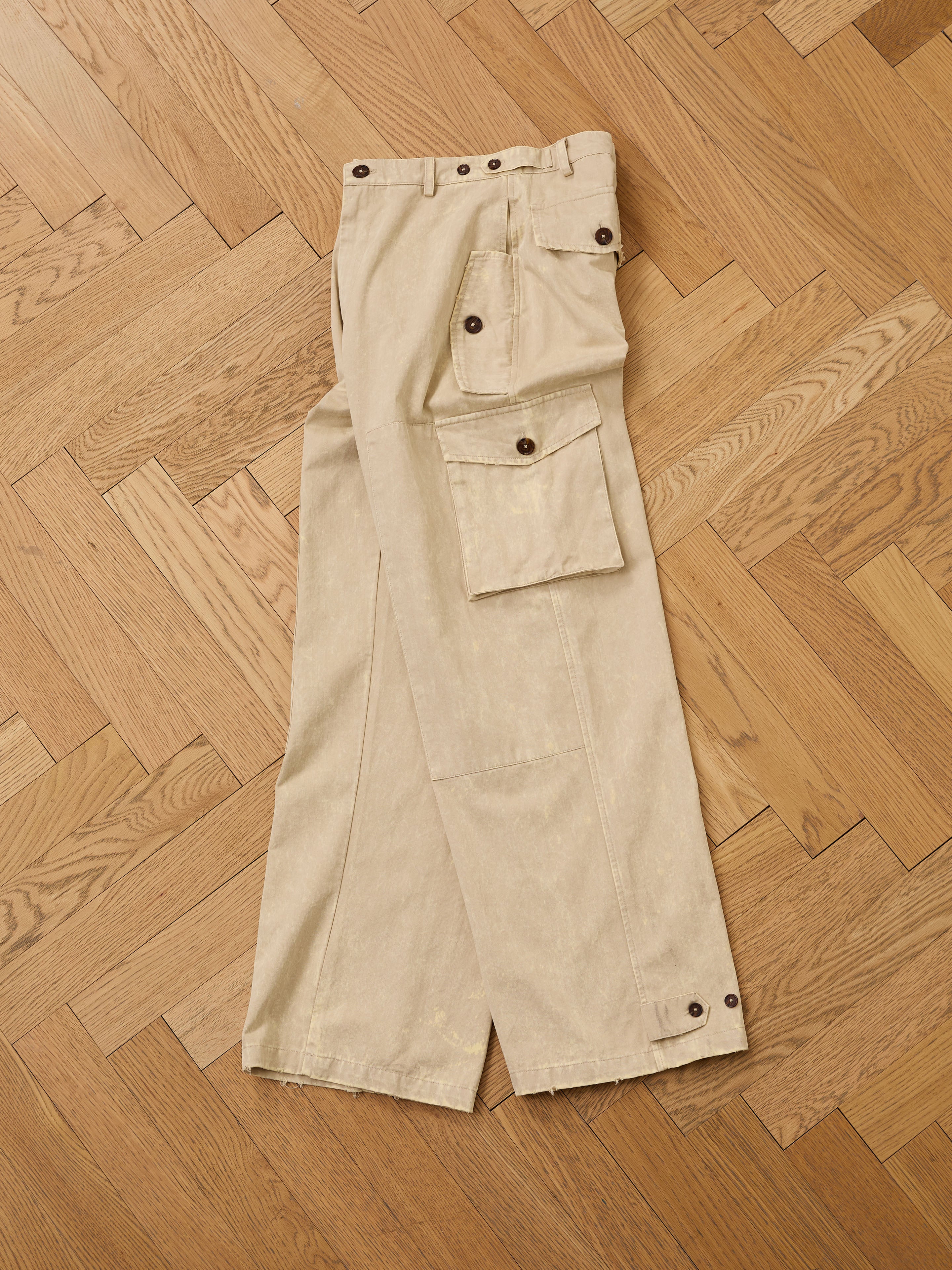 FOUND Utility Herringbone Paneled Pants in beige with large side pockets and dark buttons, unisex relaxed fit, shown laid flat on a chevron-patterned wooden floor.