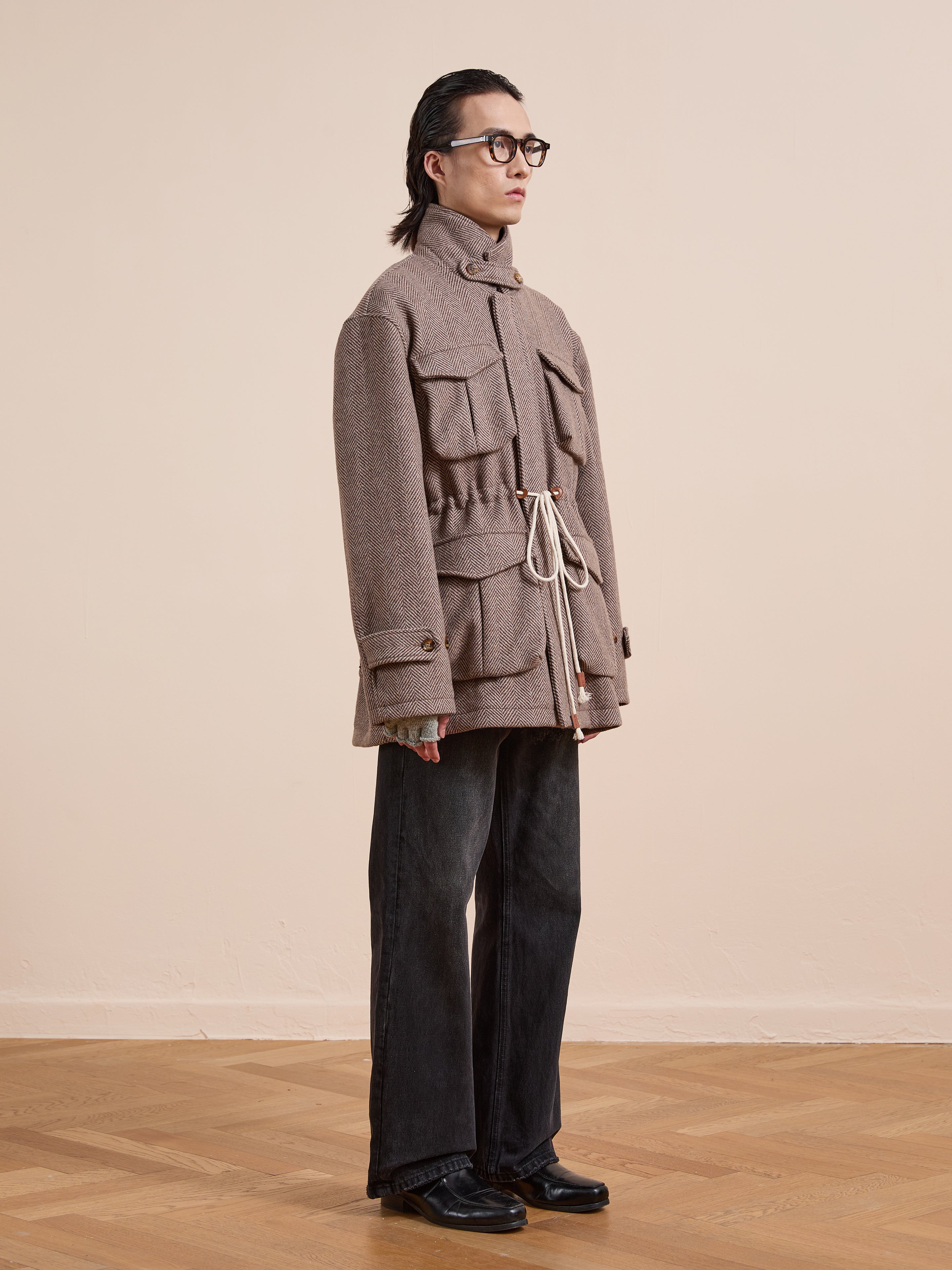 Person wearing glasses, the FOUND Herringbone Wool Barn Jacket with large pockets and a unisex relaxed fit, dark wide-leg pants, and black shoes, standing indoors on a wooden floor against a plain light wall.
