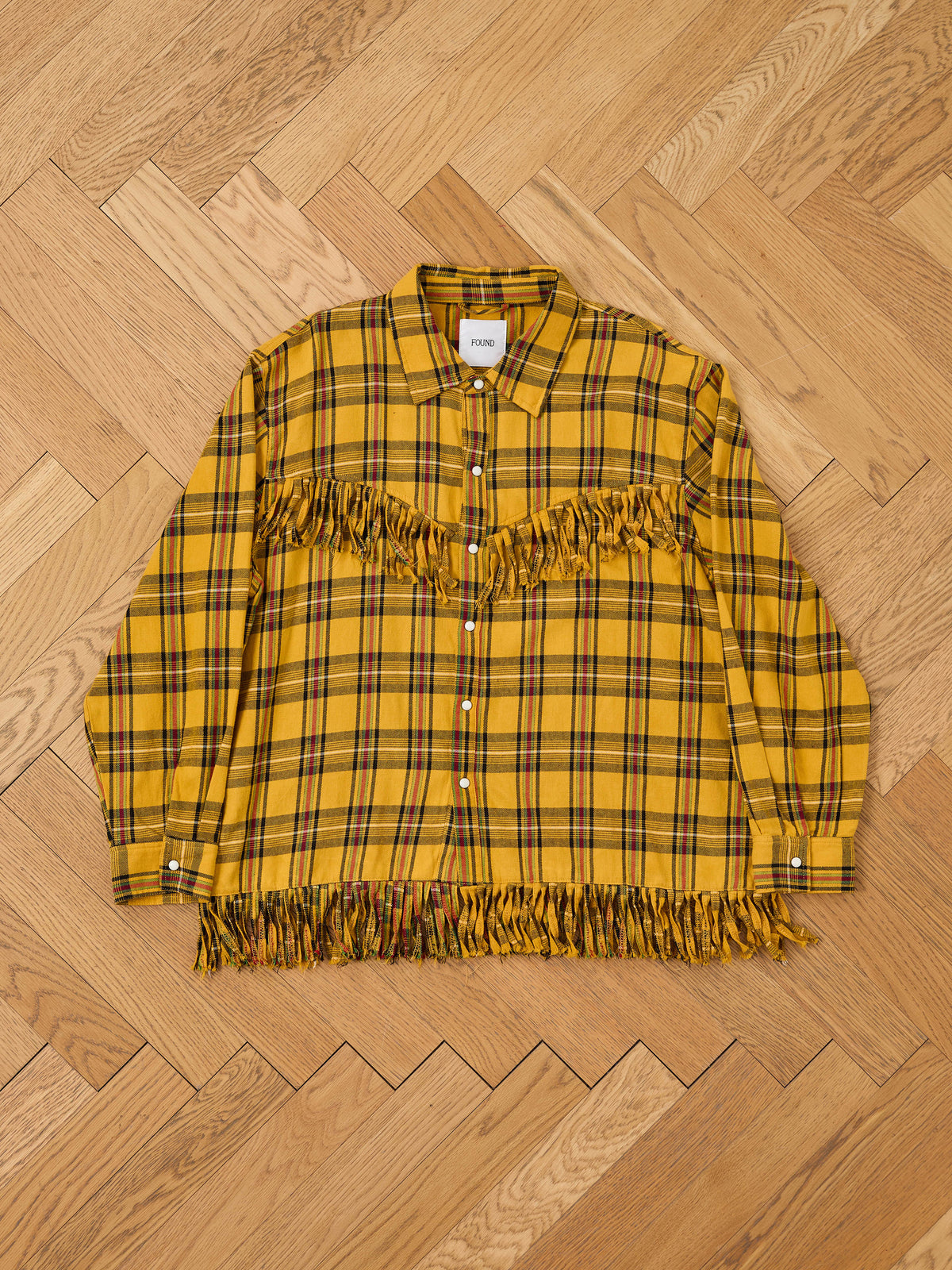 FOUND High Plains Fringe Shirt, yellow plaid unisex style with Western fringe on chest and hem, displayed flat on a light wood herringbone floor.