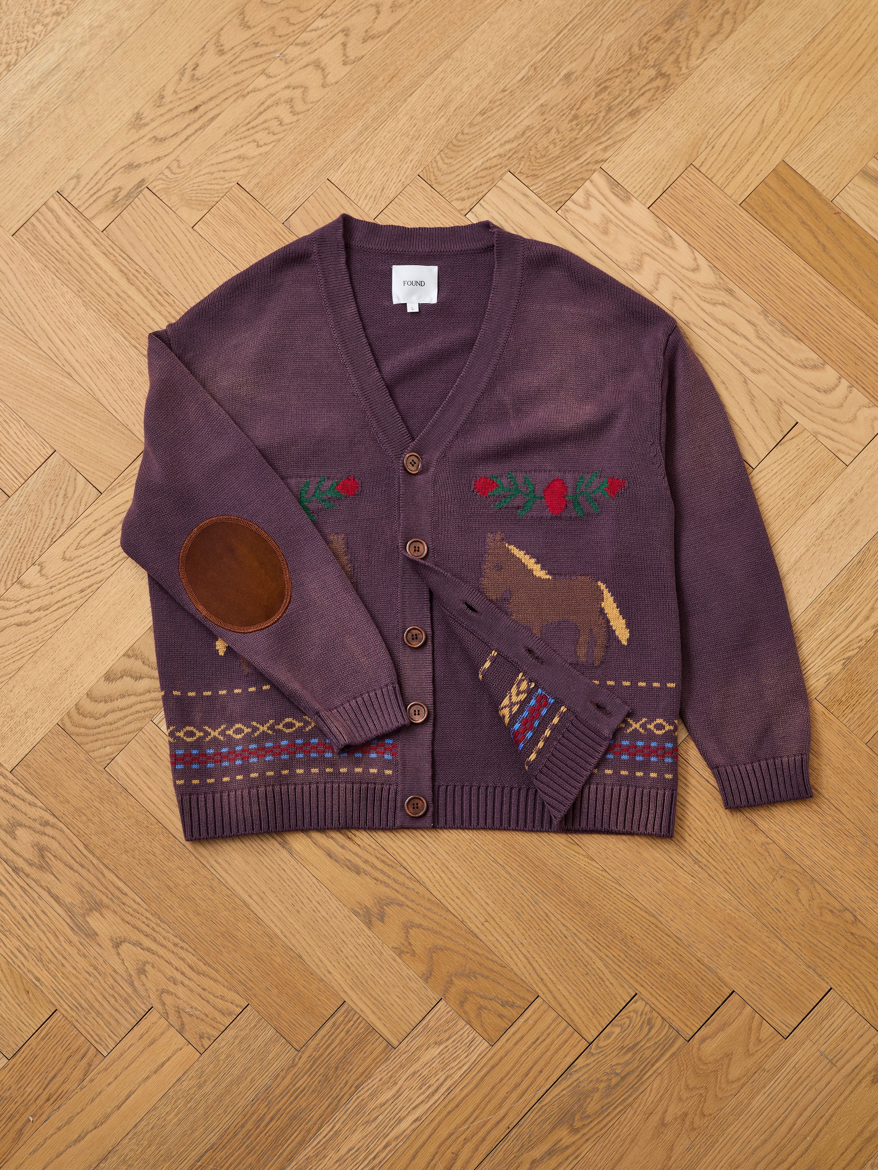 The Found Horse Jacquard Cardigan, featuring brown buttons, leather elbow patches, and horse motifs with floral and geometric patterns, is displayed flat on a wooden herringbone floor.