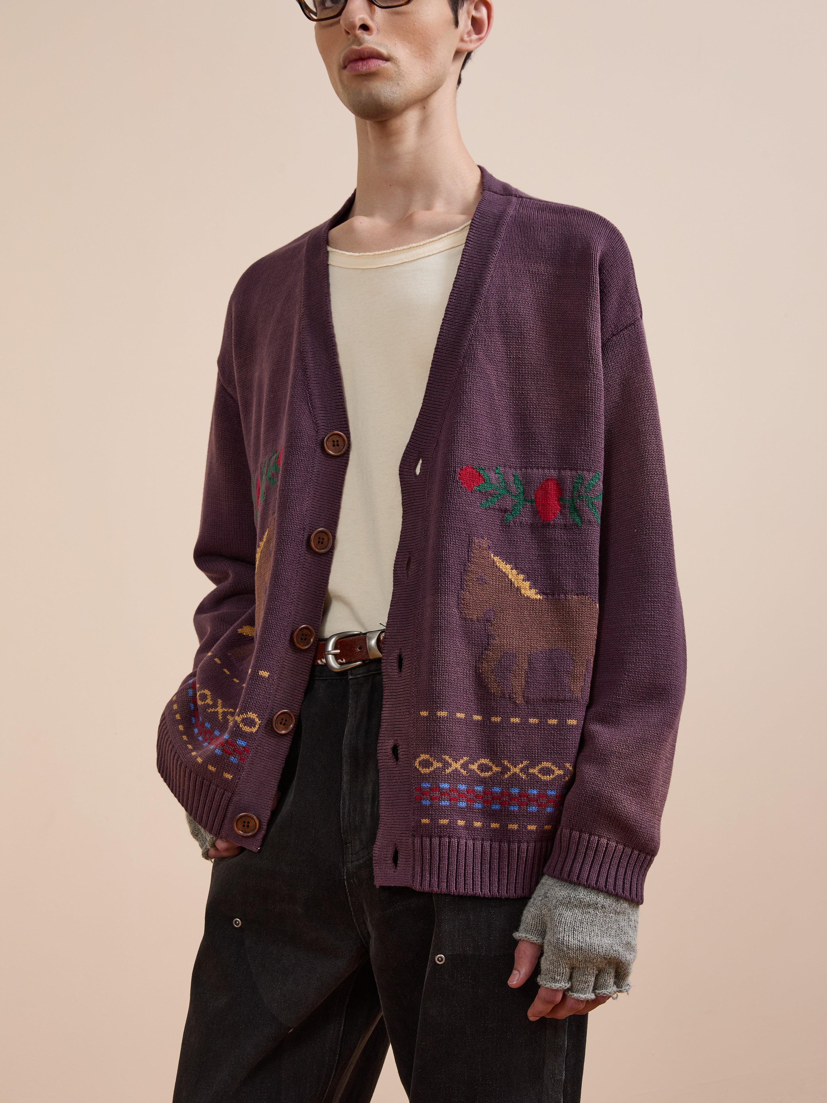 A person in Found’s Horse Jacquard Cardigan—a unisex dark purple knit with animal and floral motifs—pairs it with a cream t-shirt, black pants, grey fingerless gloves, and glasses while standing against a beige background.
