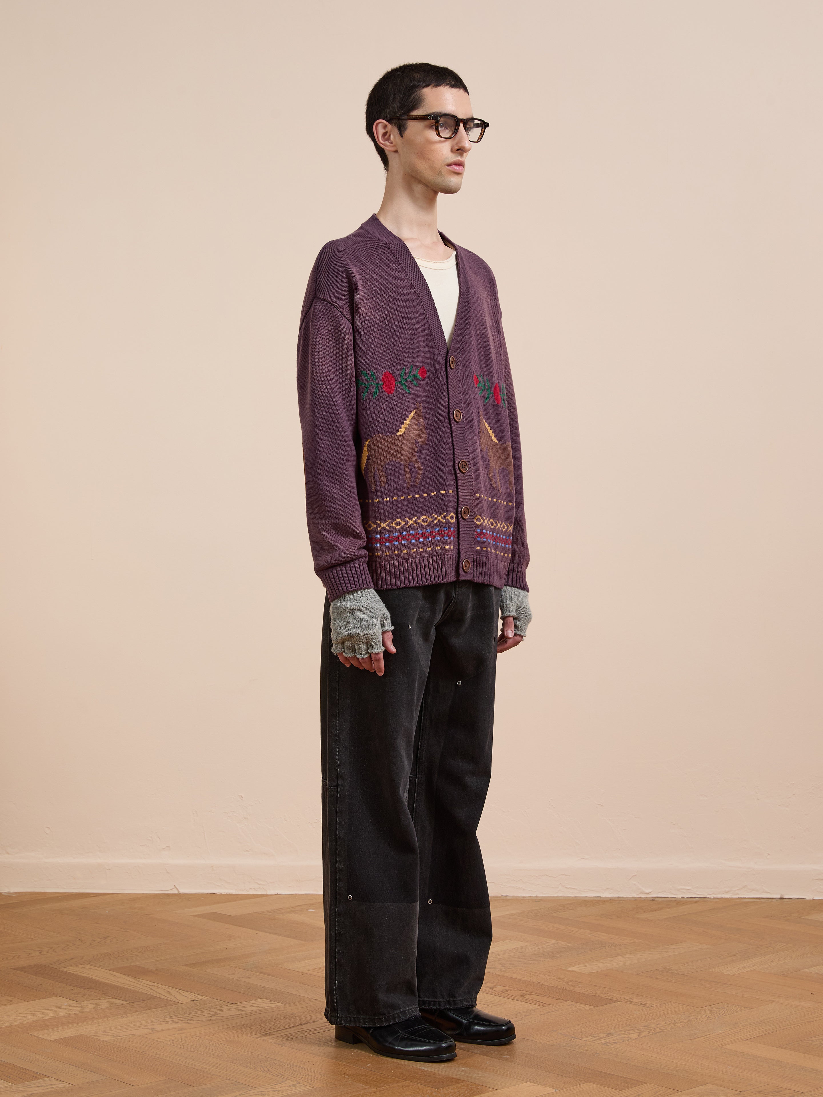 A person in Found's Horse Jacquard Cardigan, black glasses, fingerless gray gloves, black wide-leg pants, and black shoes stands on a wooden floor against a beige wall.
