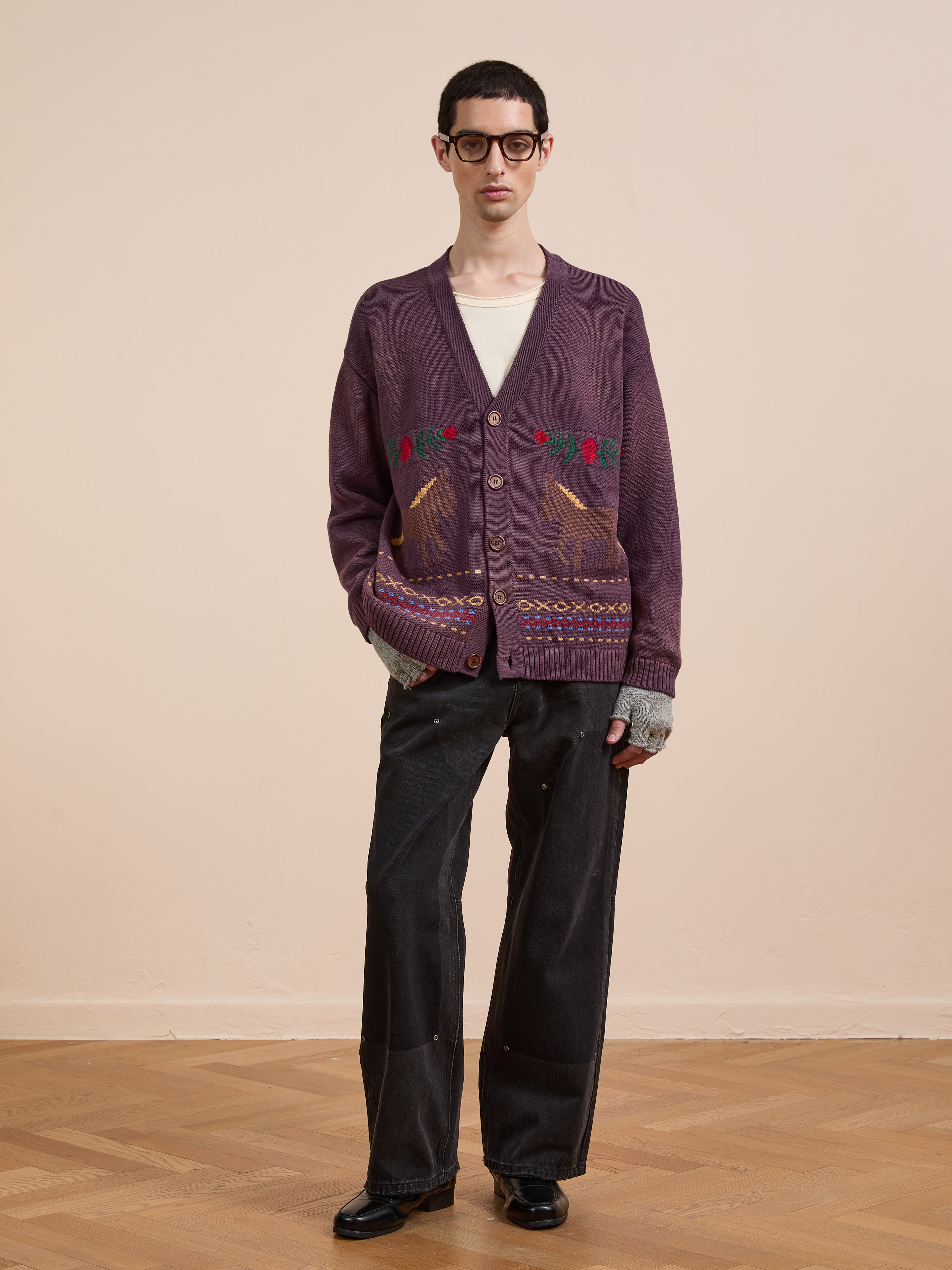A man stands indoors on a wooden floor wearing black glasses, a white T-shirt, loose dark pants, black shoes, and Found’s Horse Jacquard Cardigan in purple.