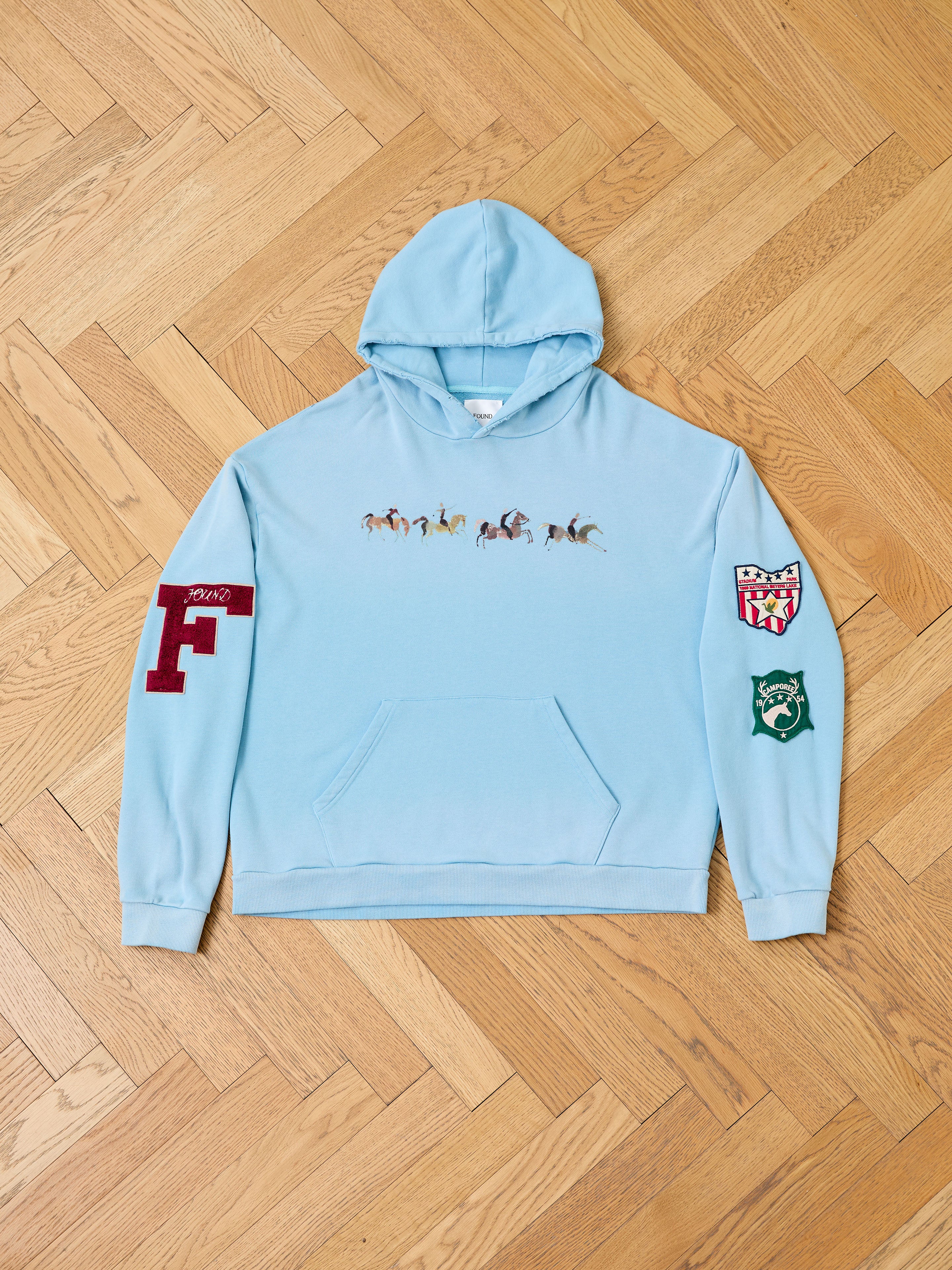 The FOUND Horse Line Hoodie in light blue, crafted from soft French Terry cotton, displays a group of small horses on the front and two patches on the sleeves, shown laid flat on a wooden floor.
