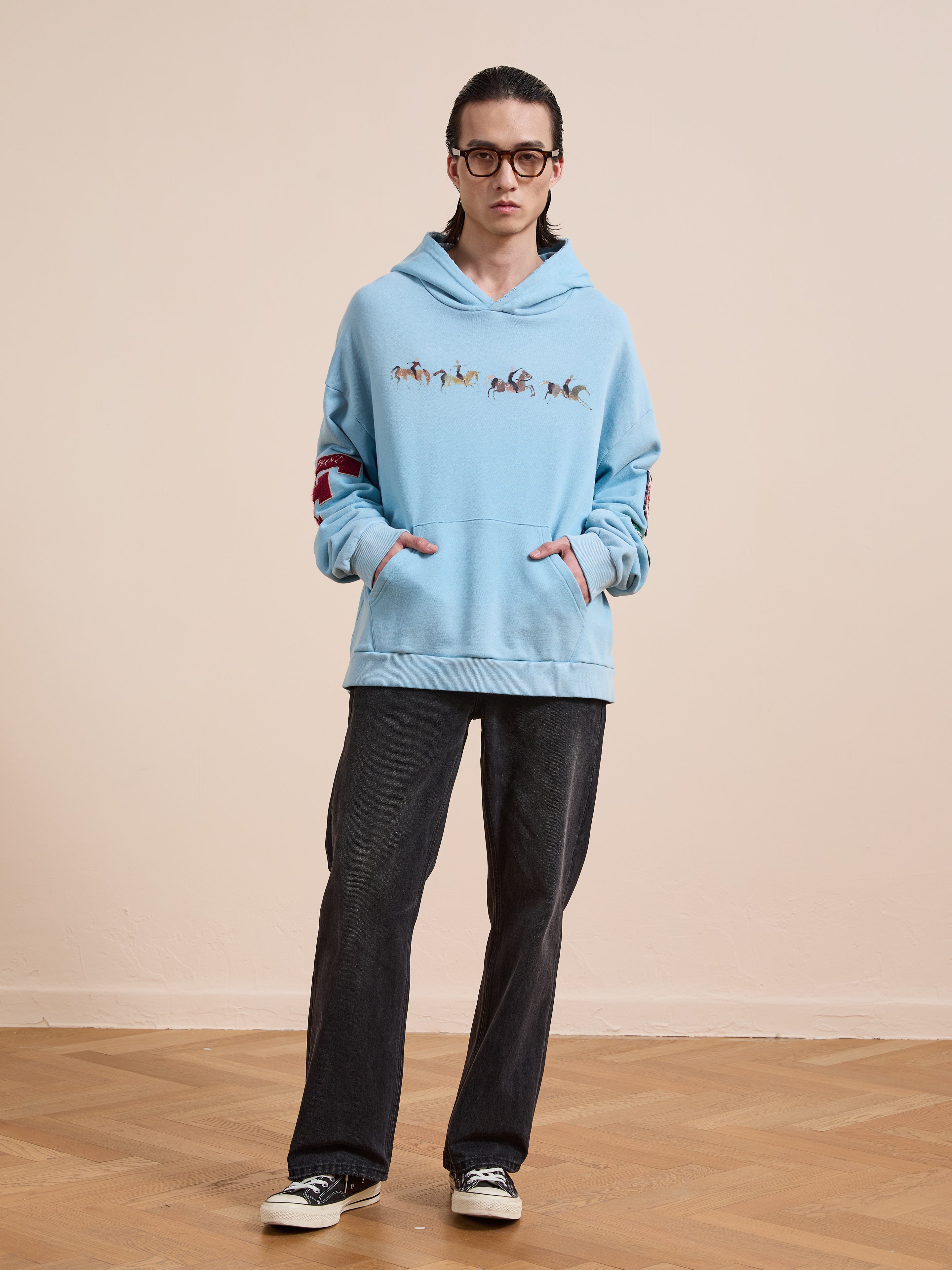A person stands indoors on a wooden floor by a plain light wall, wearing glasses, dark jeans, black sneakers, and a light blue FOUND Horse Line Hoodie crafted from soft French Terry cotton.