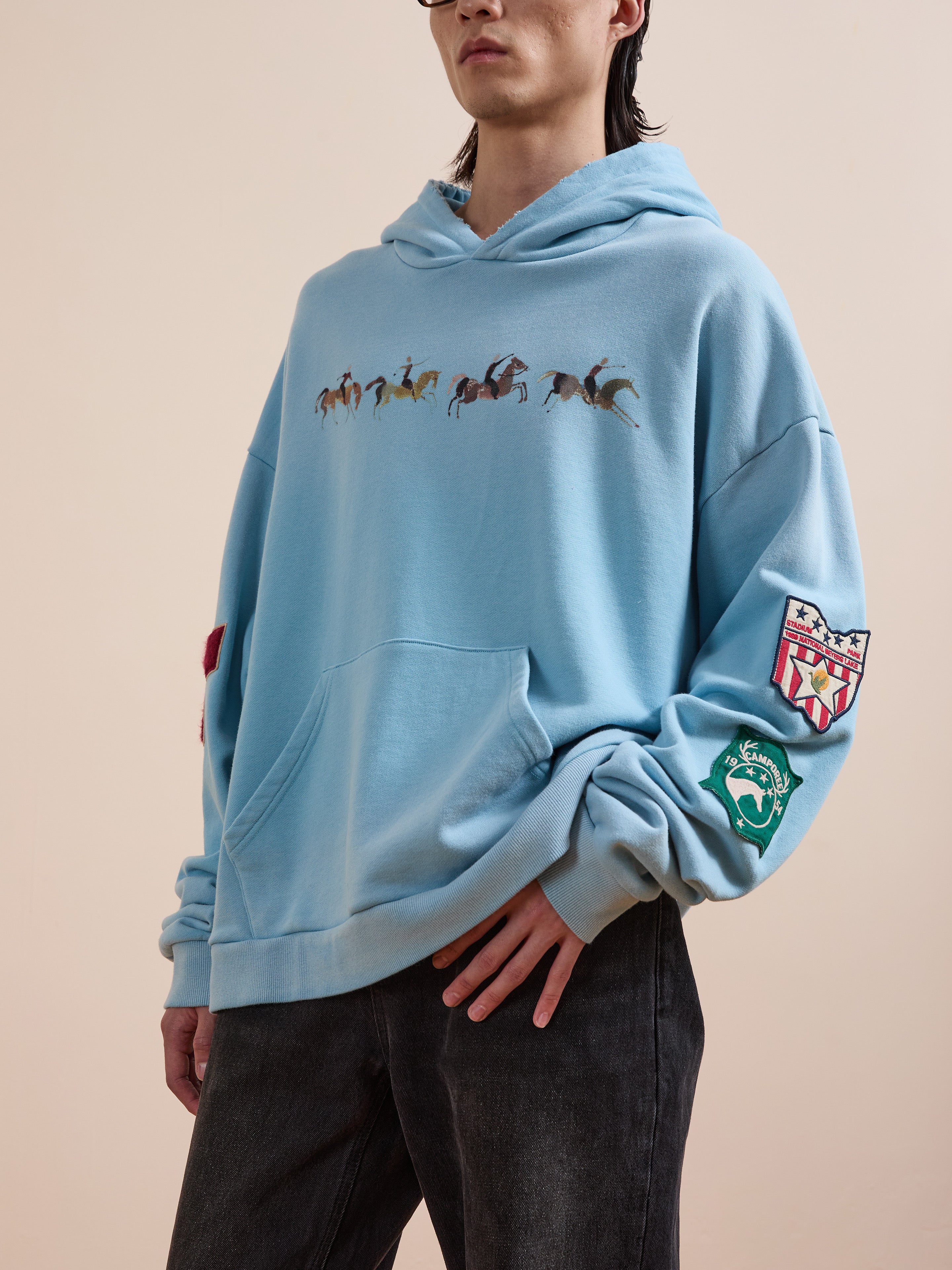 A person wears the FOUND Horse Line Hoodie, crafted from soft French Terry cotton with horse graphics on the chest and colorful sleeve patches, styled with dark jeans for a vintage look.