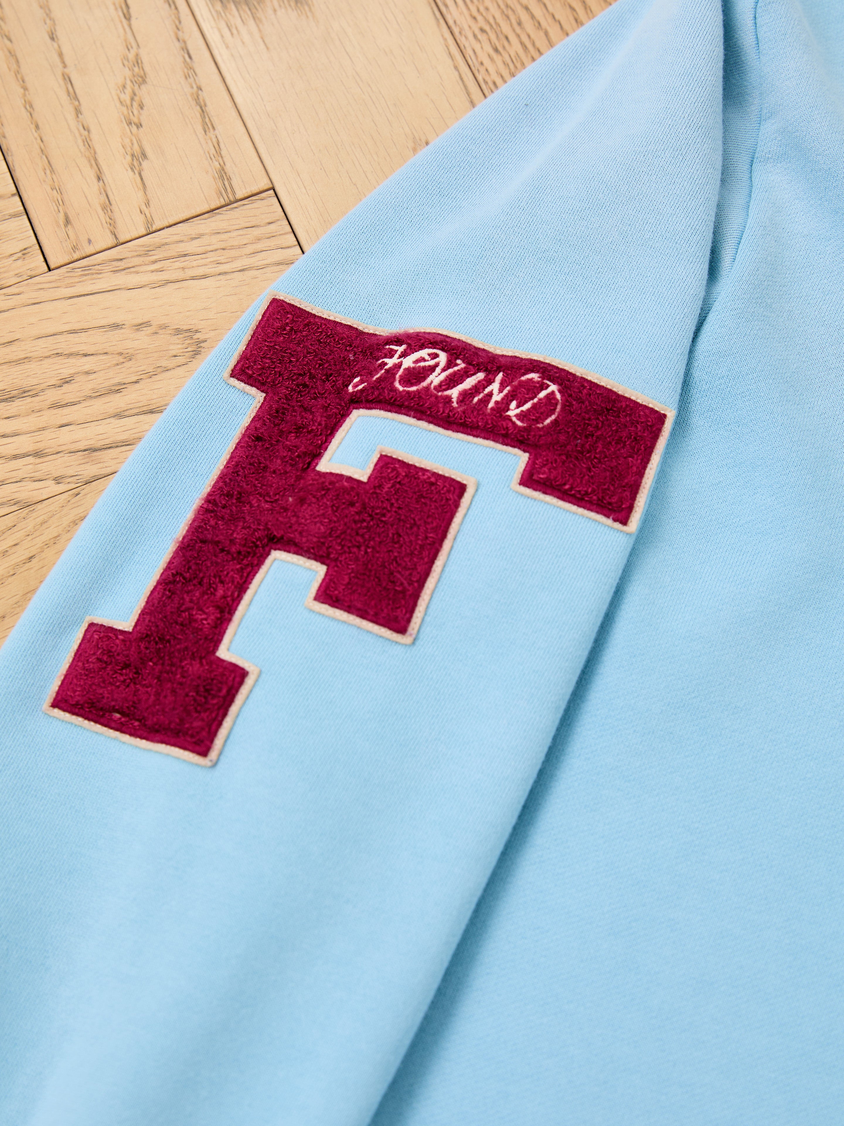 The Horse Line Hoodie by FOUND is a light blue vintage-style hoodie in soft French Terry cotton, with a bold red "F" patch and "FOUND" embroidered in white above it, displayed on a wooden surface.