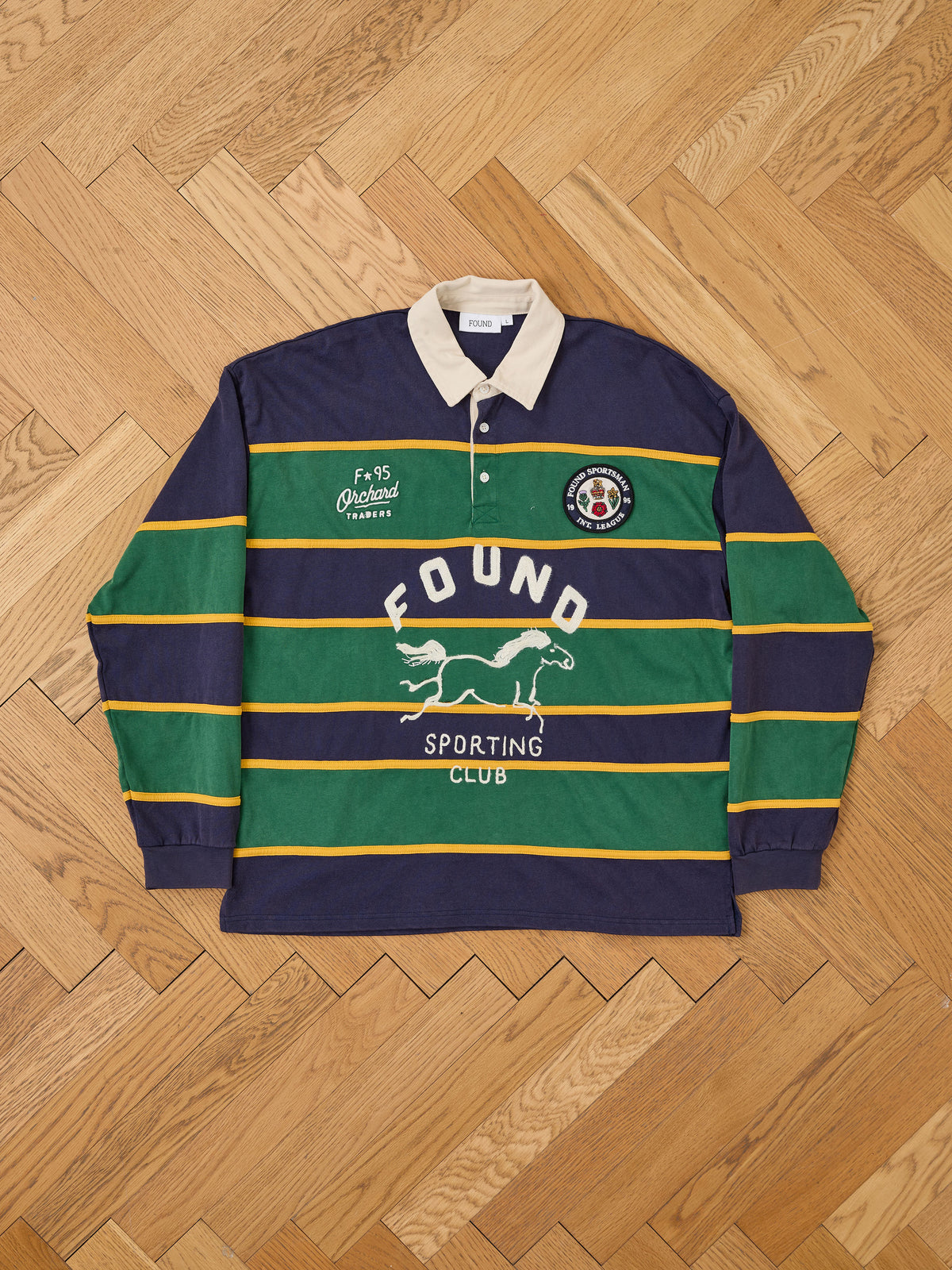 The FOUND Horse Logo Rugby is a unisex shirt with green, navy, and yellow stripes, a white collar, and “FOUND SPORTING CLUB” plus horse logo on the front.