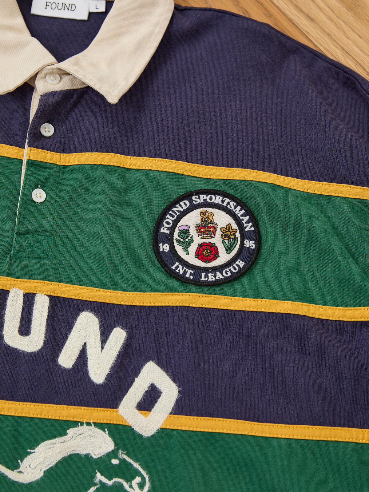 The Horse Logo Rugby by FOUND is a unisex shirt with a beige collar, embroidered crest patch, and partial “FOUND” lettering displayed on a wooden surface.