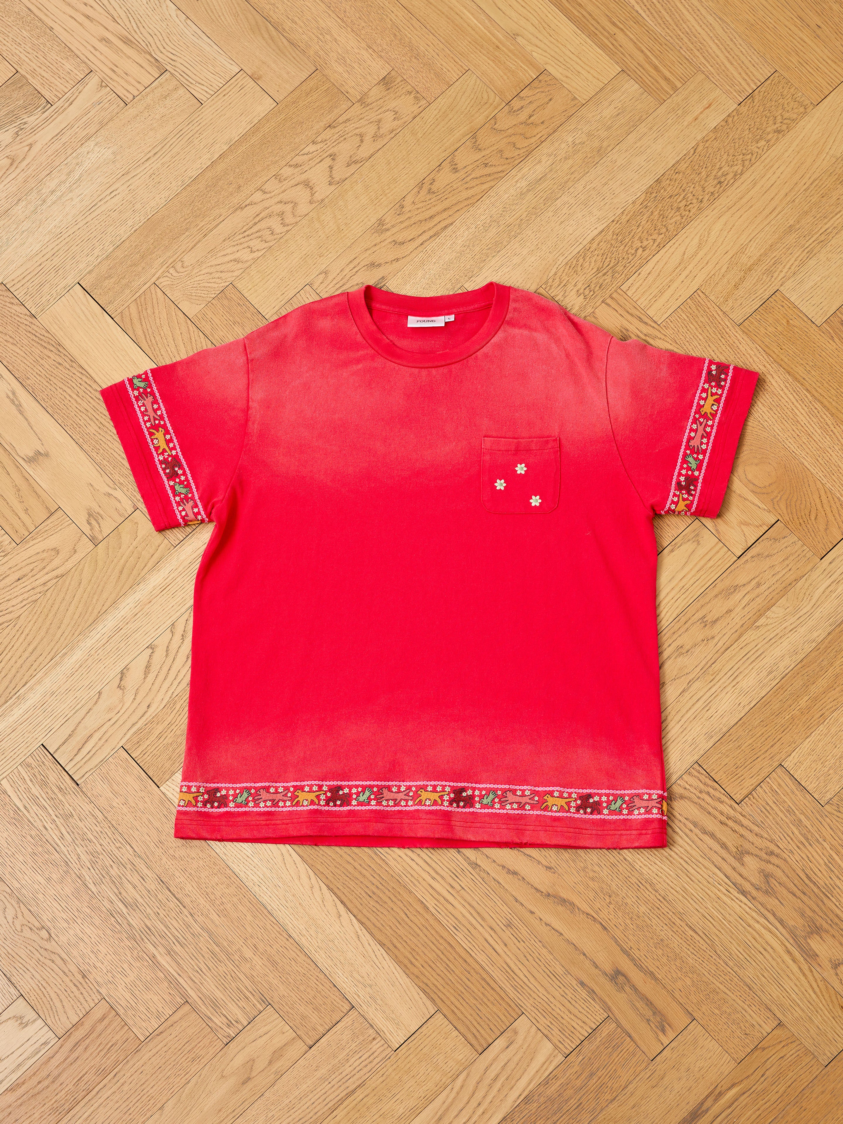 The FOUND Horse Motif Embroidered Tee is a unisex relaxed fit red vintage wash short-sleeve shirt with a chest pocket and patterned sleeve and hem bands, displayed flat on a wooden herringbone floor.
