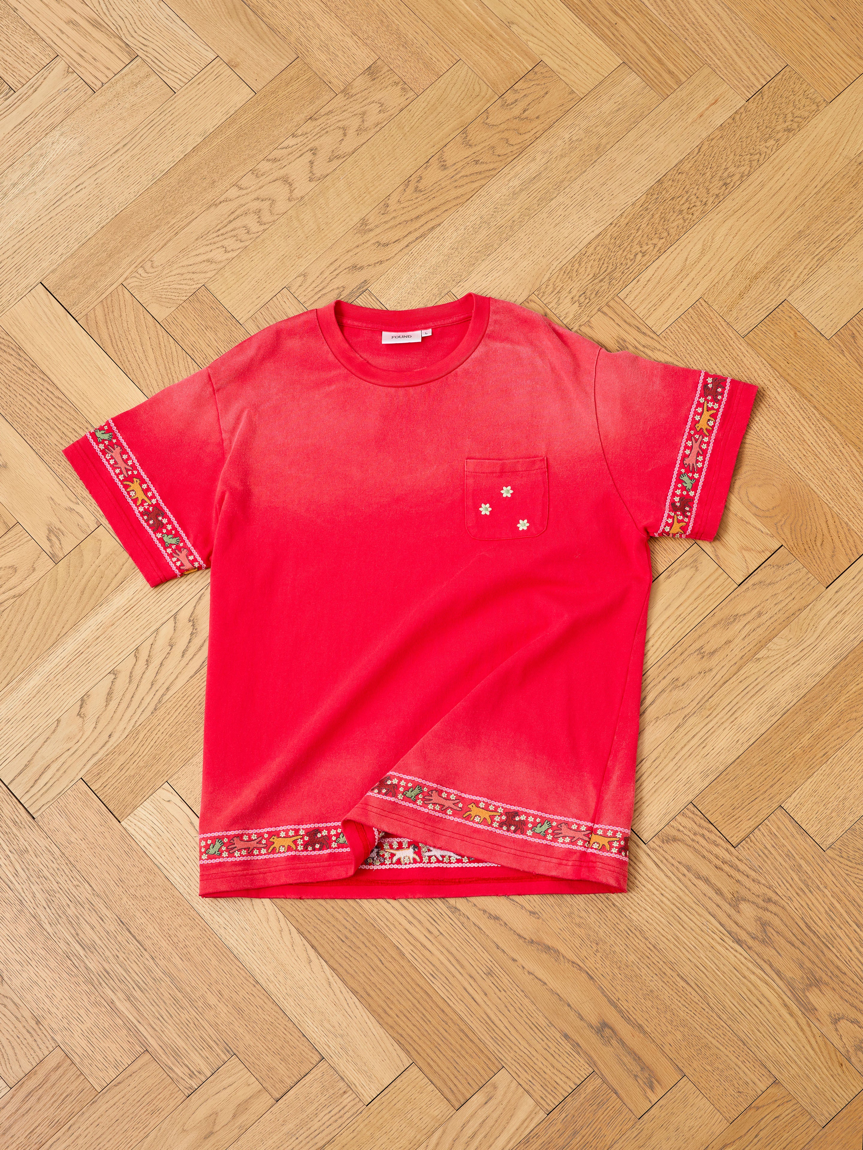 The FOUND Horse Motif Embroidered Tee is a red unisex relaxed fit short-sleeve shirt with a chest pocket, horse embroidery on the pocket, and patterned trim on the sleeves and hem, shown laid flat on a wooden herringbone floor.
