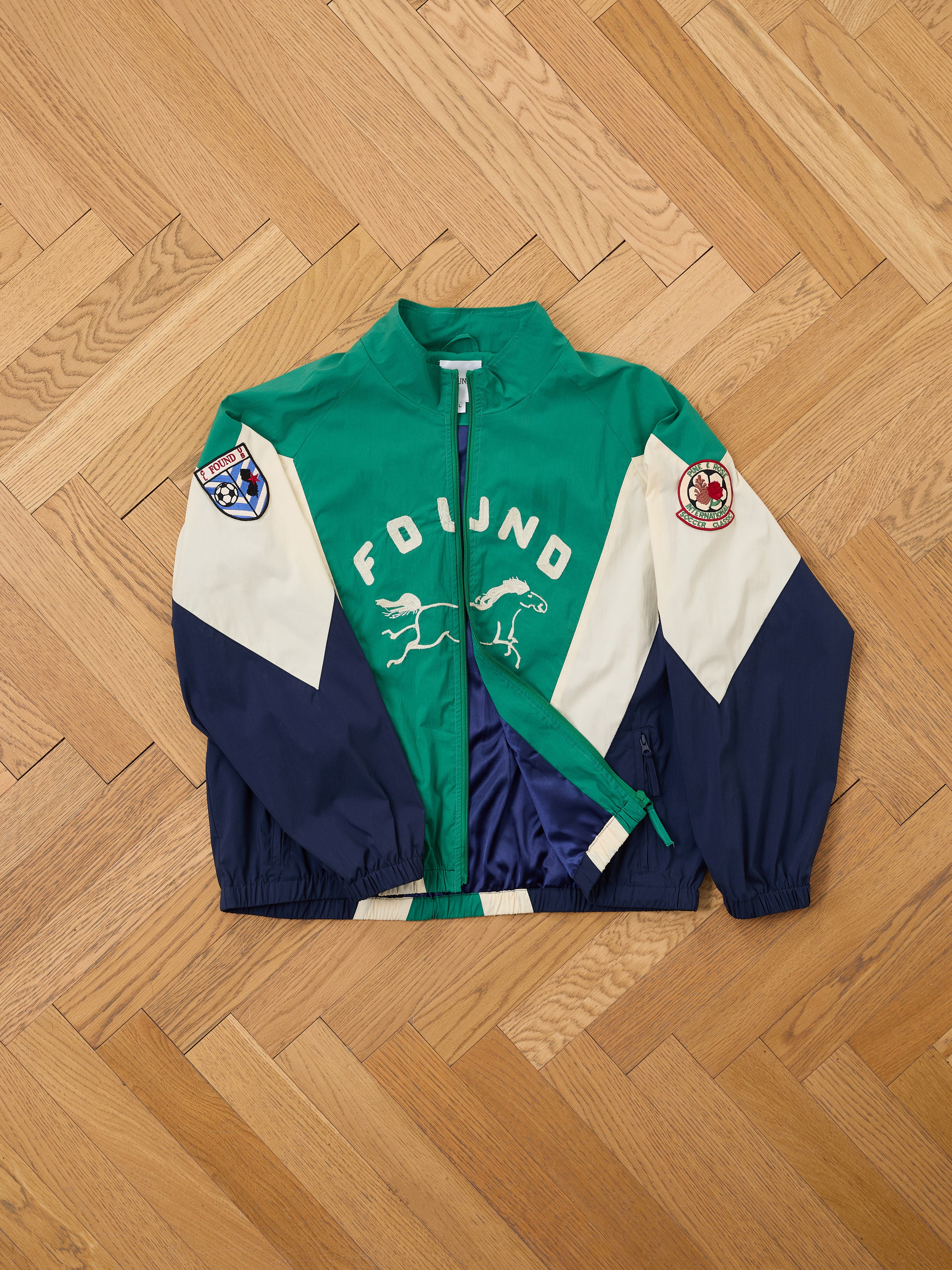 A green, navy, and white Crestline Sport Zip Jacket by FOUND with animal graphics, "FOUND" text, and two sleeve patches is laid flat on a wooden herringbone floor—a standout vintage sportswear piece.