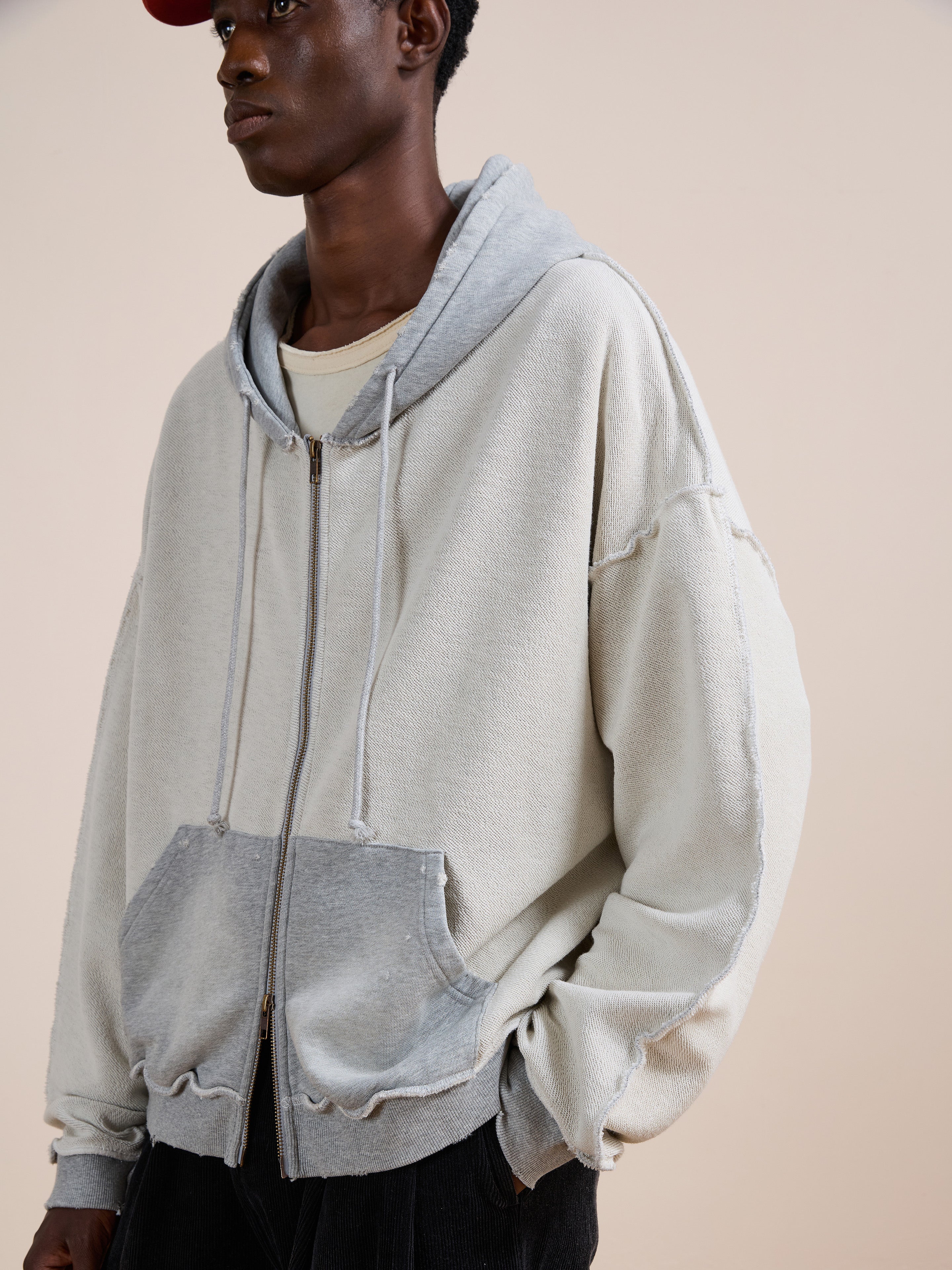A person wearing FOUND's Deconstructed Terry Zip Up Hoodie in light gray, featuring a front pocket and drawstrings, layered over a cream shirt with a red cap, standing against a plain background.