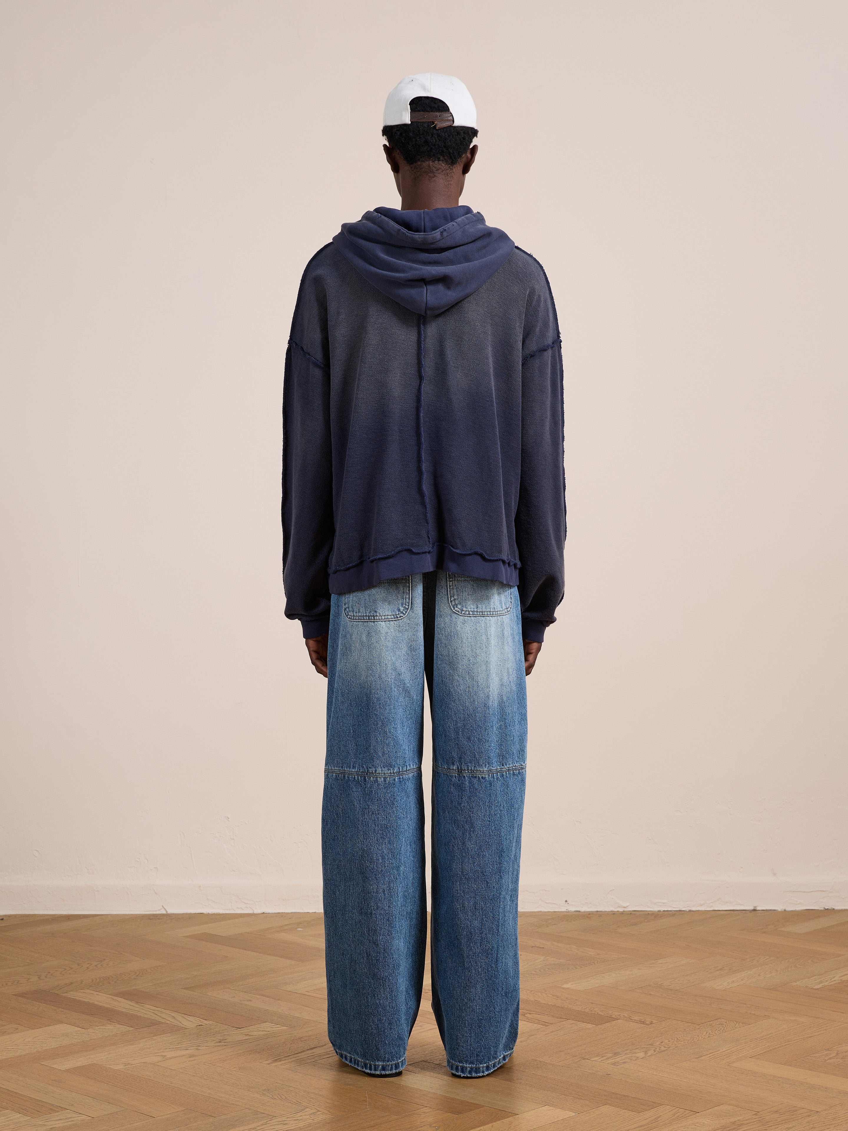 Indoors, a person faces away wearing FOUND's navy Deconstructed Terry Zip Up Hoodie with a relaxed fit, wide-leg blue jeans, and a white cap. The floor is wooden and the wall is plain beige.