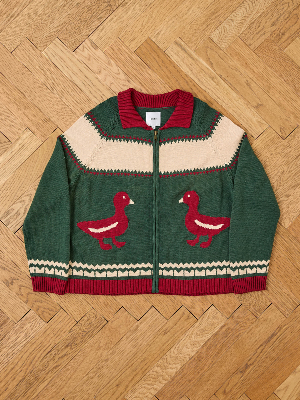 The FOUND Isles Sweater is a vintage green knit cardigan with red trim, beige accents, and red duck designs, shown flat on a wooden floor.