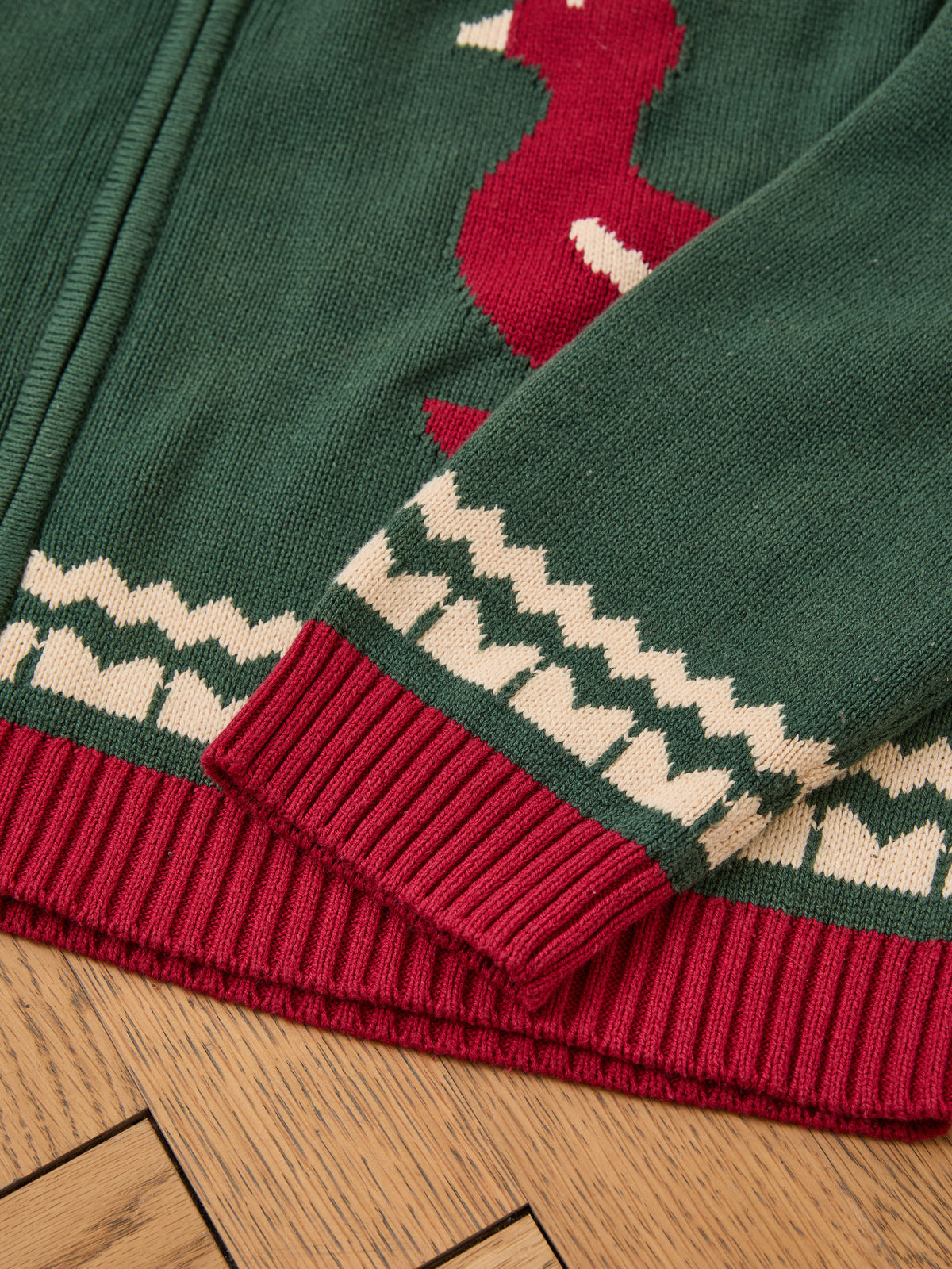 Close-up of the FOUND Isles Sweater, green vintage knitwear with red and white bird pattern and red ribbed cuffs, displayed on a wooden floor.