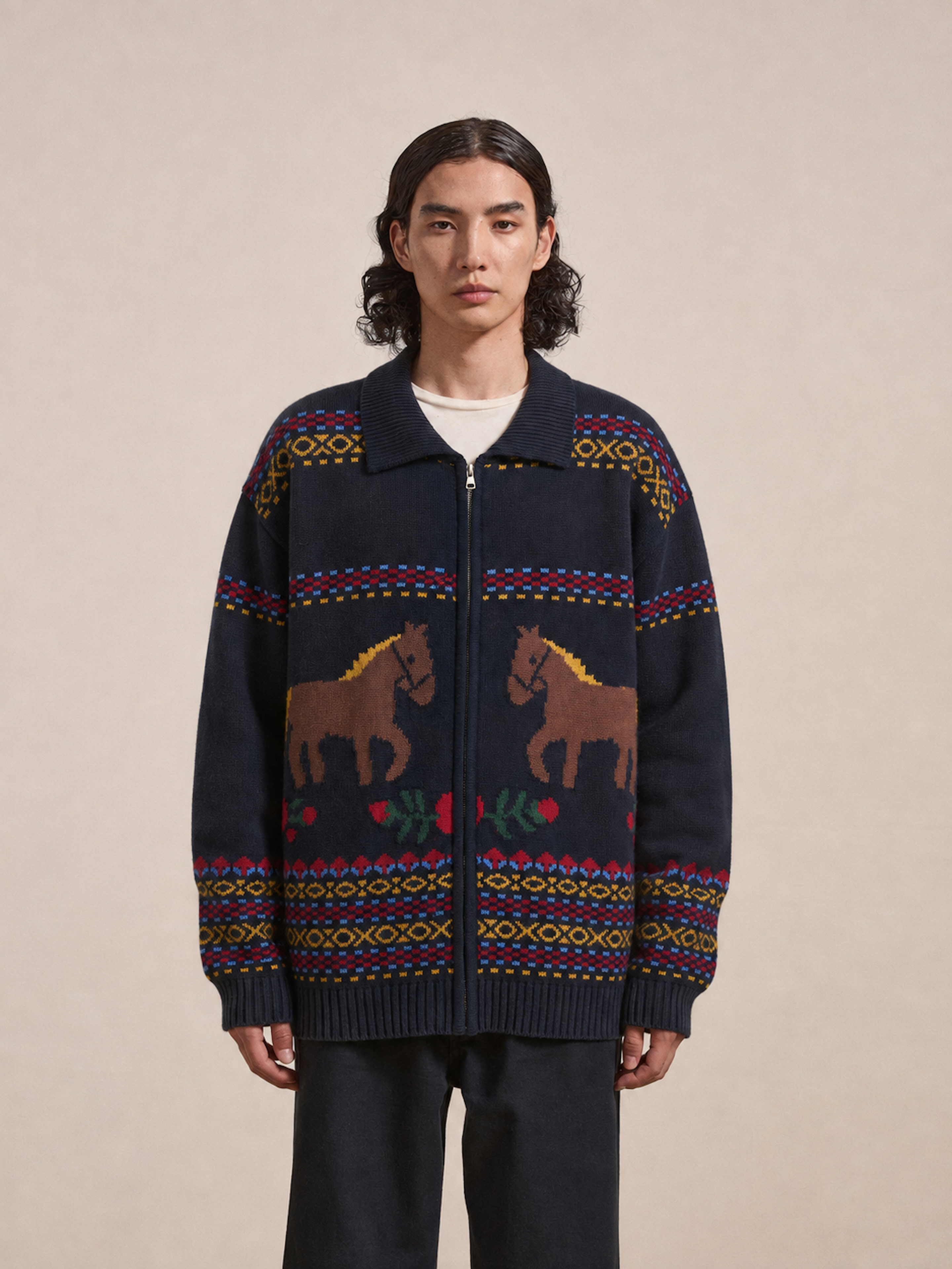 A person stands facing forward, wearing the Isles Sweater by Found—a navy knit zip cardigan detailed with jacquard horse motifs and floral patterns, accented by geometric designs in red, yellow, and green.