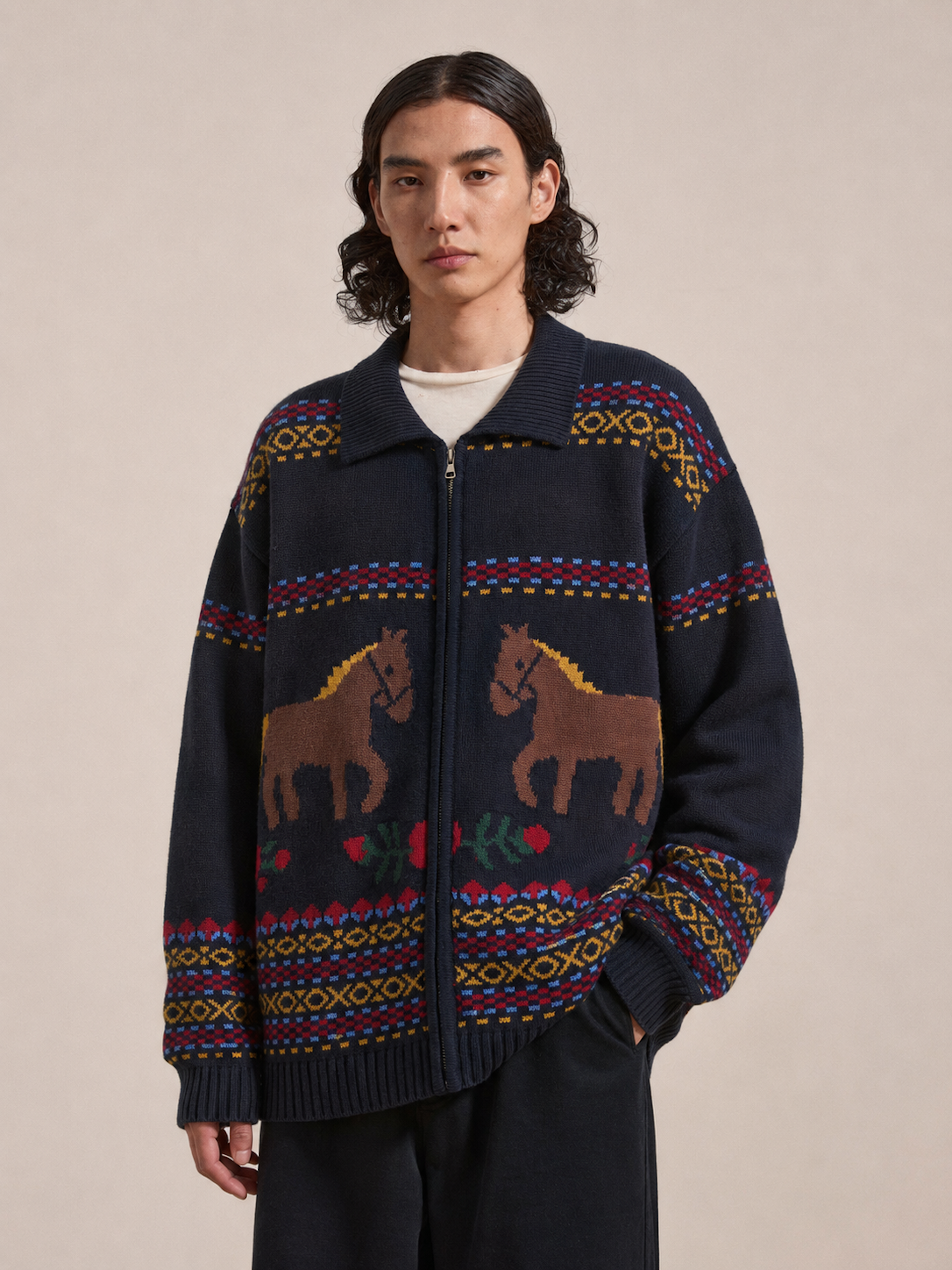 A person wearing the Found Isles Sweater—a navy knit zip cardigan adorned with colorful jacquard horse motifs—stands against a plain beige background.
