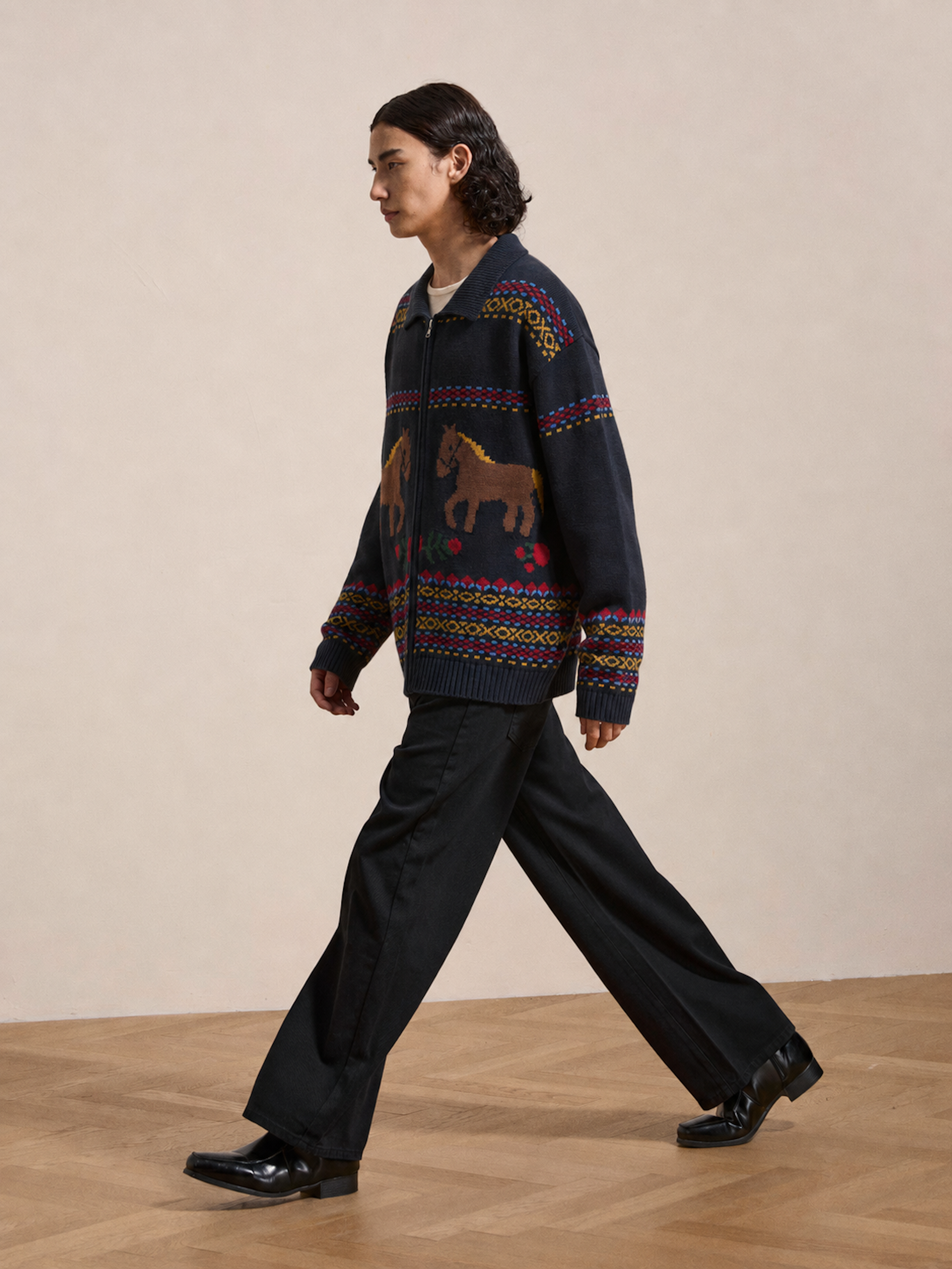 A person walks indoors on a wooden floor, wearing black pants, black shoes, and the Isles Sweater by Found—a navy knit zip cardigan featuring colorful jacquard horse motifs.