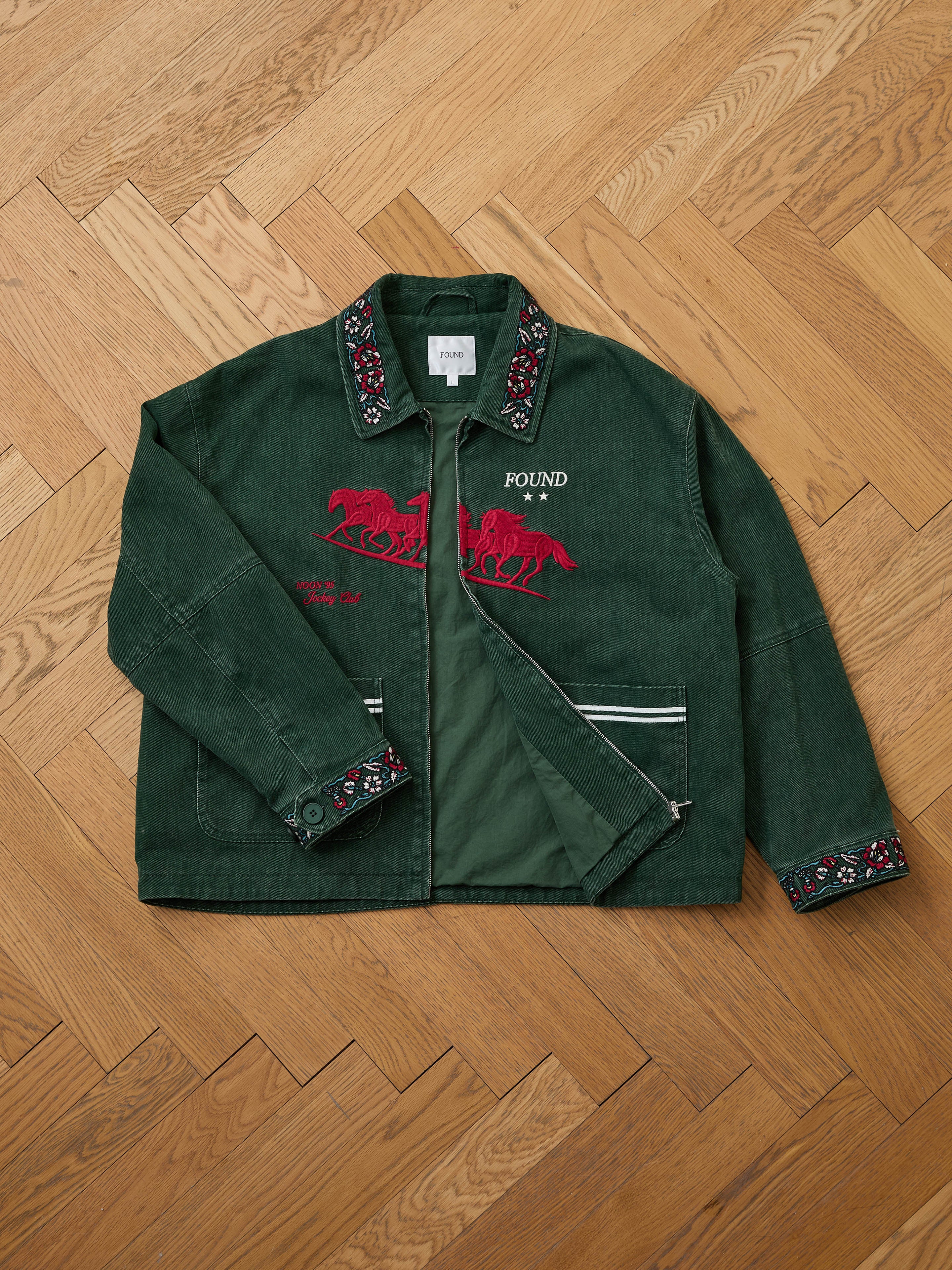 The Found Jockey Club Embroidered Jacket is a unisex relaxed fit piece with racing horses embroidery, "FOUND" text, and decorative collar and cuffs, shown on a wooden herringbone floor.