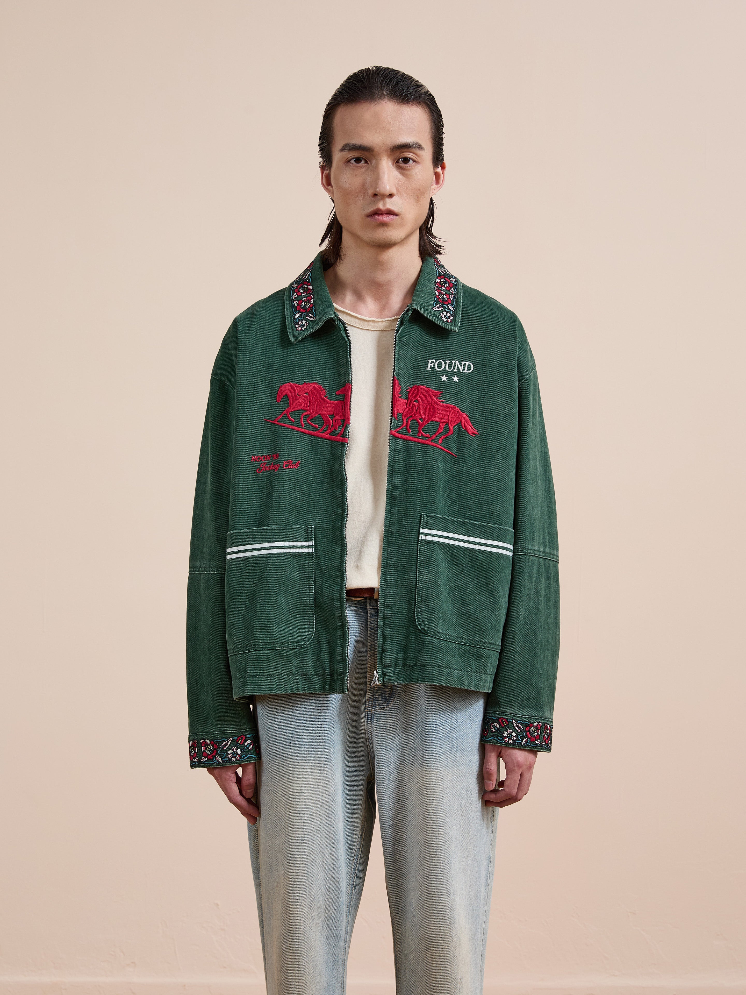 A person with long hair faces forward, wearing the Found Jockey Club Embroidered Jacket in green over a white shirt and light blue jeans. The unisex relaxed fit and racing horses embroidery offer casual comfort against a beige background.