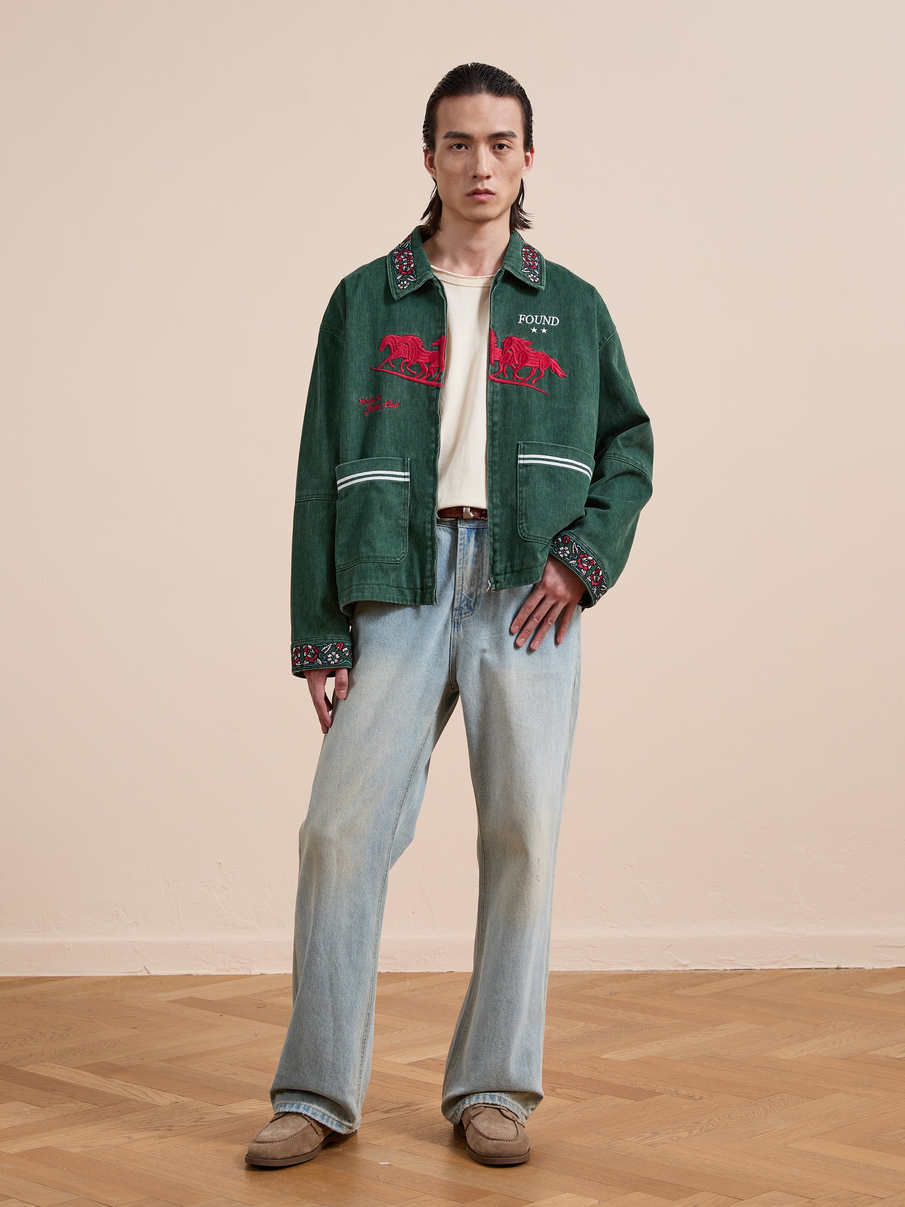 Indoors on a wooden floor, a person in a neutral pose wears Found's Jockey Club Embroidered Jacket, paired with light wash jeans, cream shirt, and beige shoes for a unisex relaxed look.