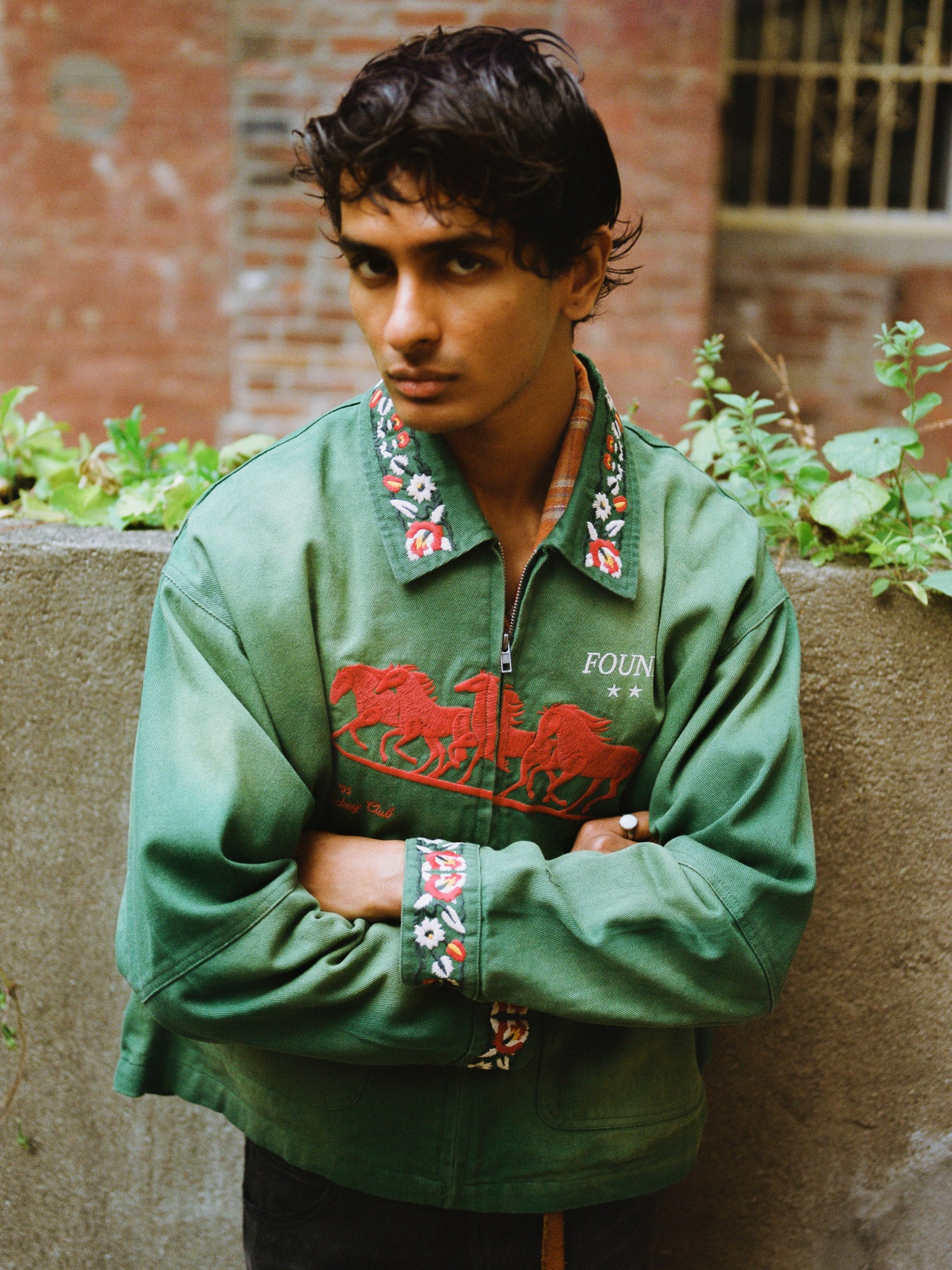 A young man with dark hair stands outdoors by a concrete wall, wearing the Found Jockey Club Embroidered Jacket in a unisex relaxed fit, looking directly at the camera with his arms crossed.