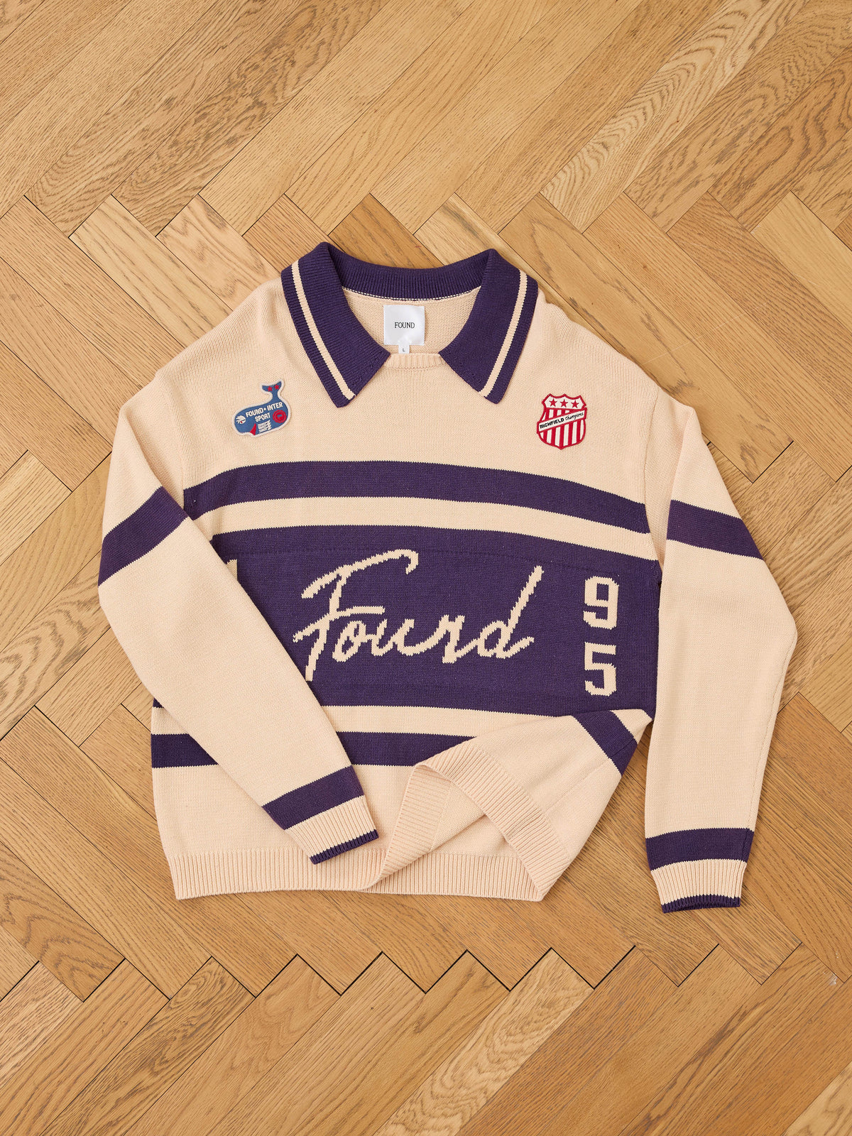 FOUND Knit Jersey in beige and purple with “Found 95” text and patches, shown laid flat on a herringbone wood floor.