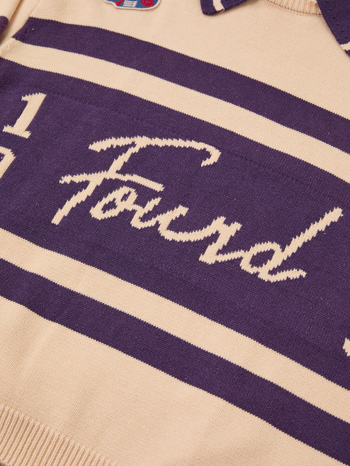 Close-up of the Knit Jersey by FOUND: beige varsity knitwear with wide purple stripes, “Found” in cursive, and a small embroidered patch near the collar.