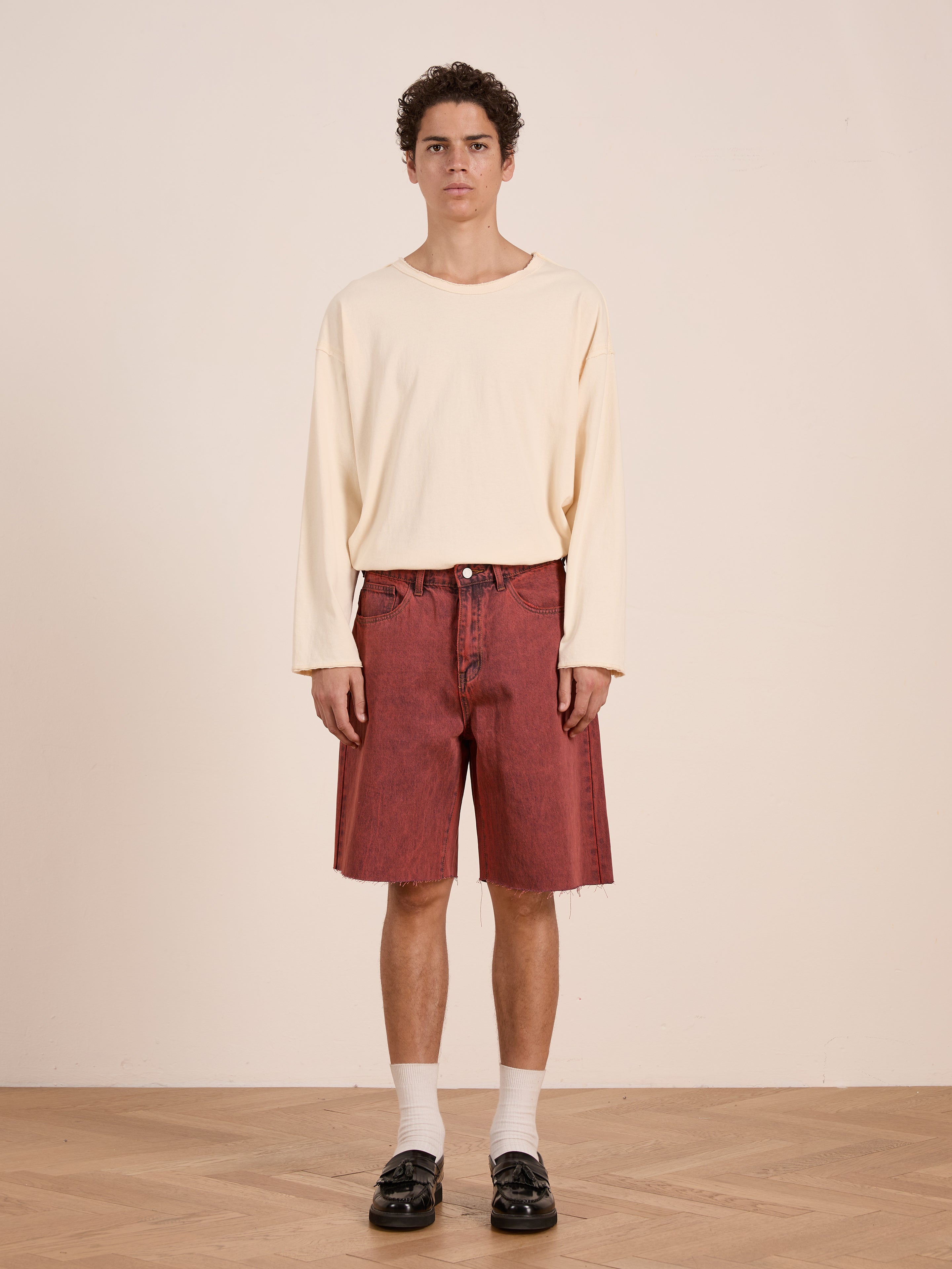 Indoors, a person wears a loose cream long-sleeve shirt, FOUND Lacy Jean Jorts in vintage red wash denim, white socks, and black loafers against a plain background.