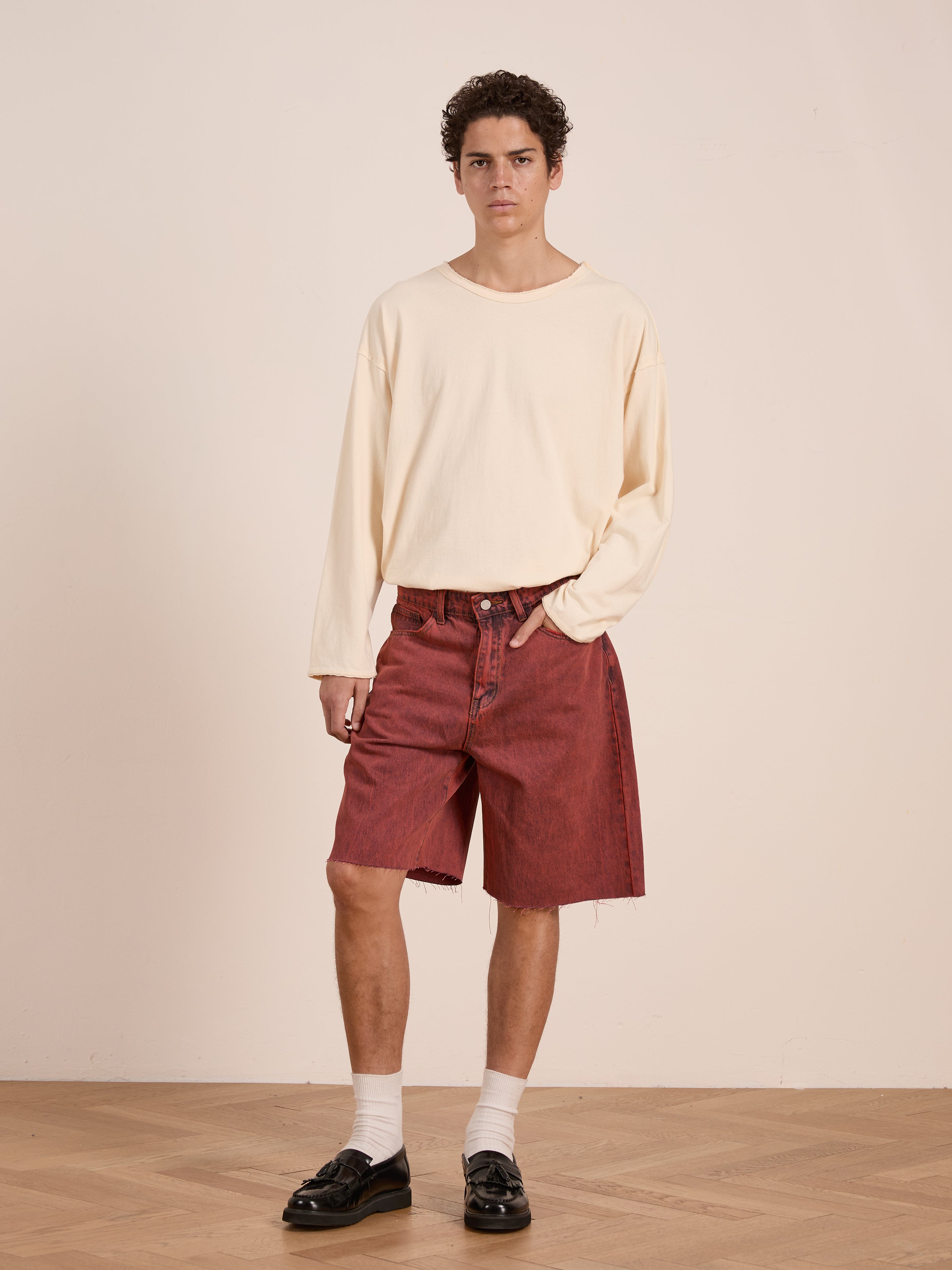 A young man stands indoors against a light wall and wooden floor, wearing FOUND's Lacy Jean Jorts in vintage red wash, paired with a cream long-sleeve shirt, white socks, and black loafers.