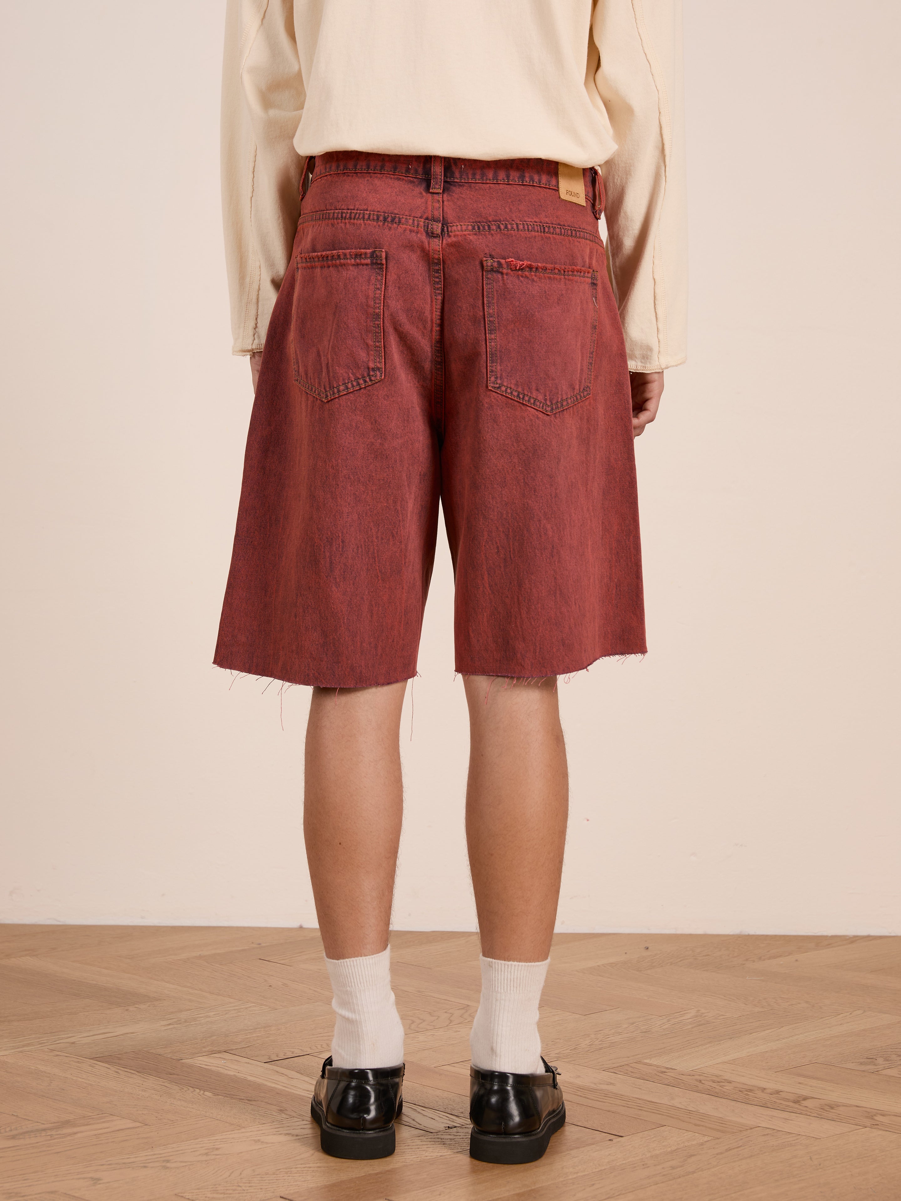 A person stands on a wooden floor facing away from the camera, wearing a cream long-sleeve shirt, FOUND Lacy Jean Jorts in vintage red wash 100% cotton denim with raw hems, white socks, and black shoes.