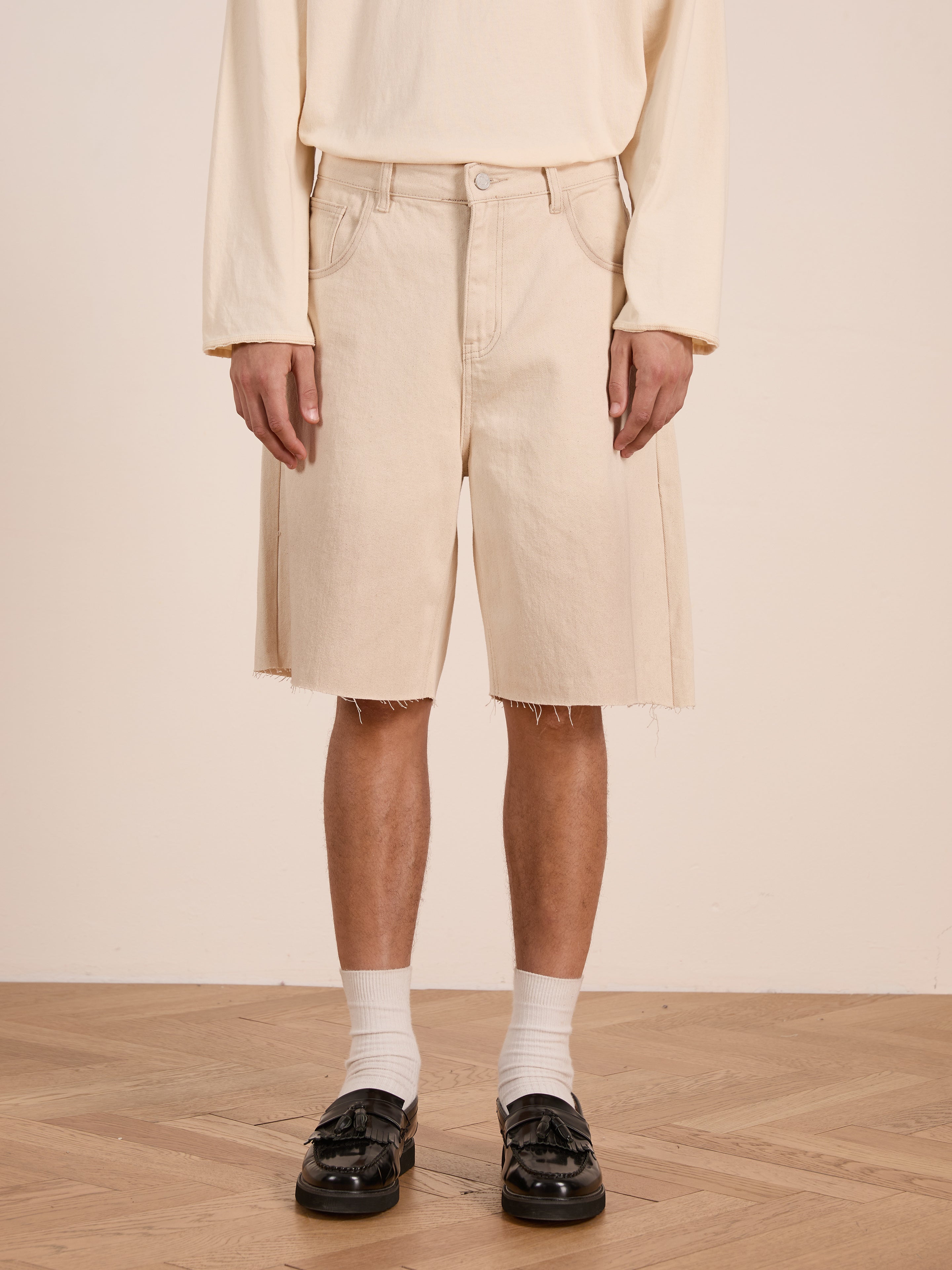 A person wearing FOUND's Lacy Jean Jorts, a long-sleeve beige shirt, white socks, and black loafers stands on a wooden floor against a plain background.