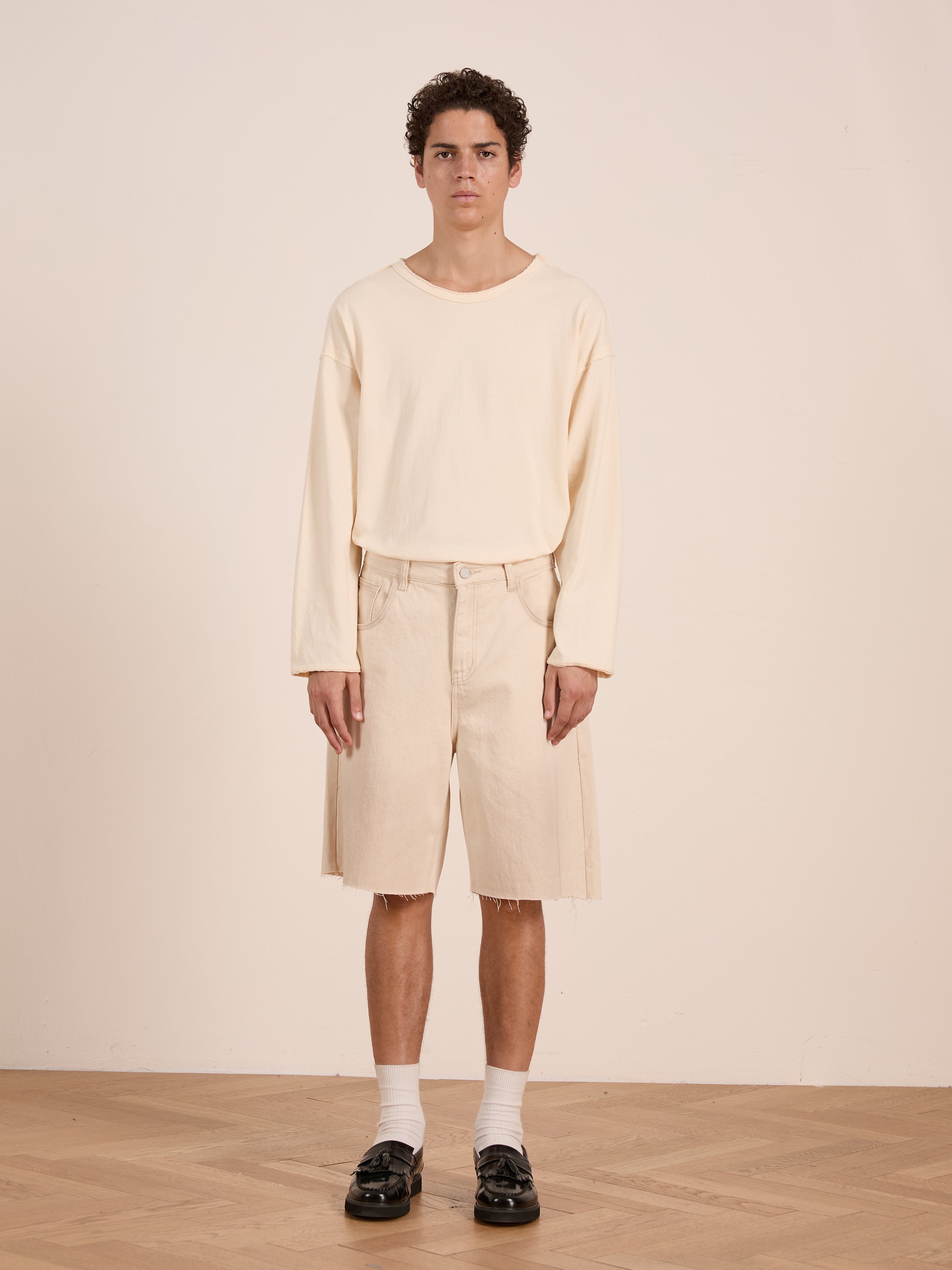 A person stands indoors against a light wall and wooden floor, wearing FOUND's Lacy Jean Jorts, a long-sleeve beige shirt, white socks, and black loafers.