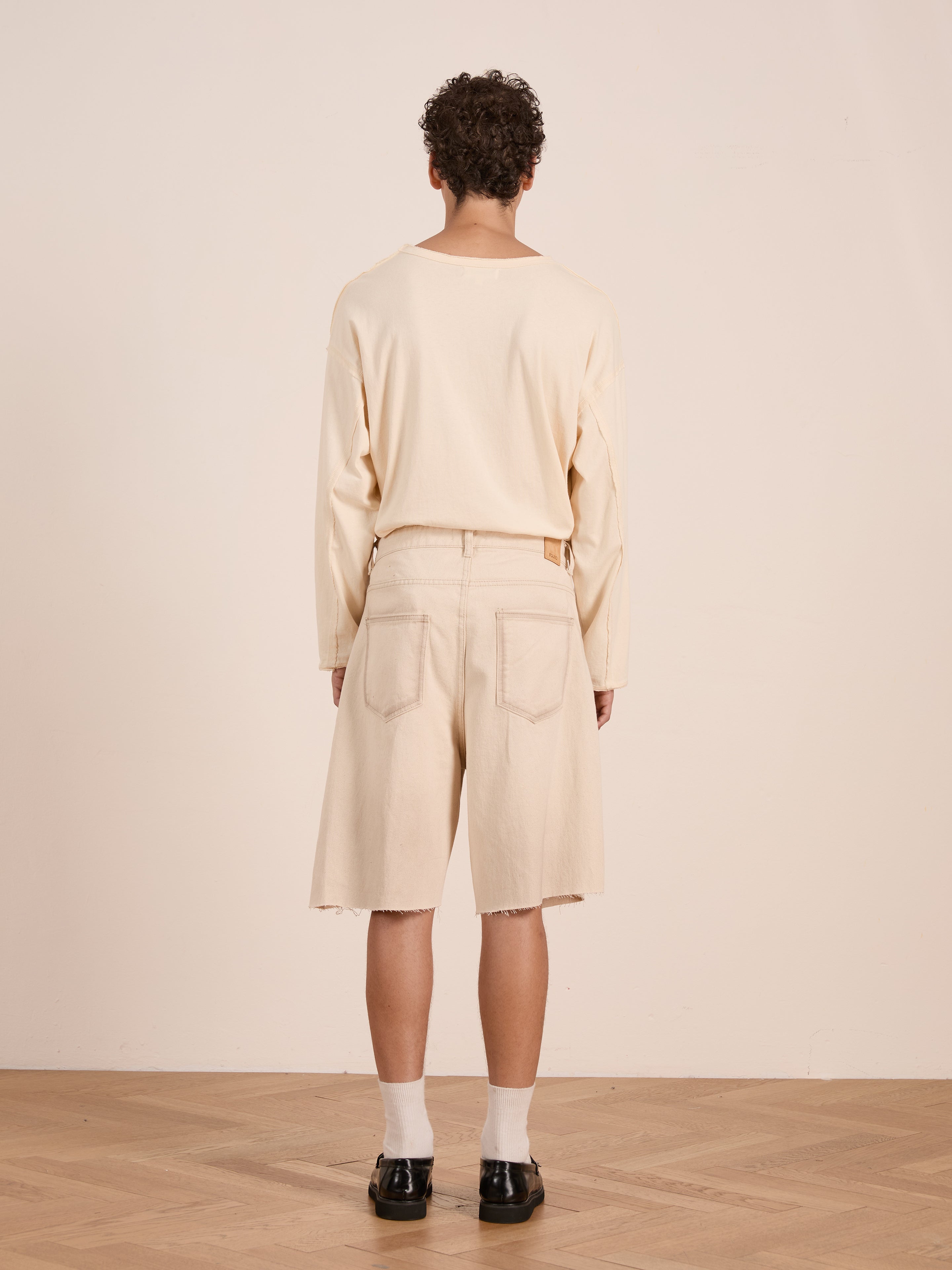 A person stands indoors with their back to the camera, wearing FOUND's Lacy Jean Jorts, a long-sleeve cream shirt, white socks, and black shoes on a wooden floor against a plain light background.