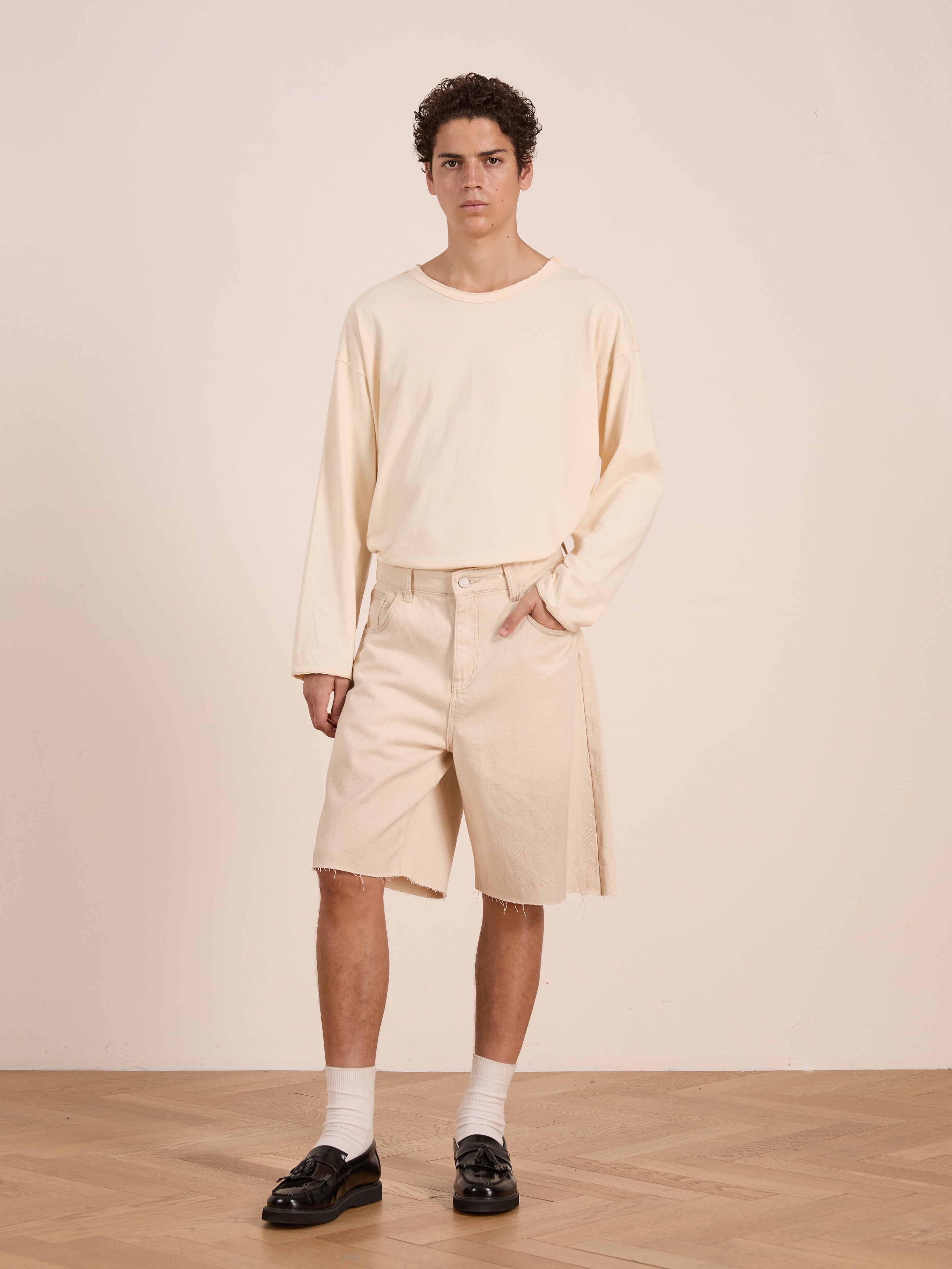 A person stands indoors against a plain light wall, wearing a loose cream long-sleeve shirt, FOUND Lacy Jean Jorts with frayed hems, white socks, and black loafers.