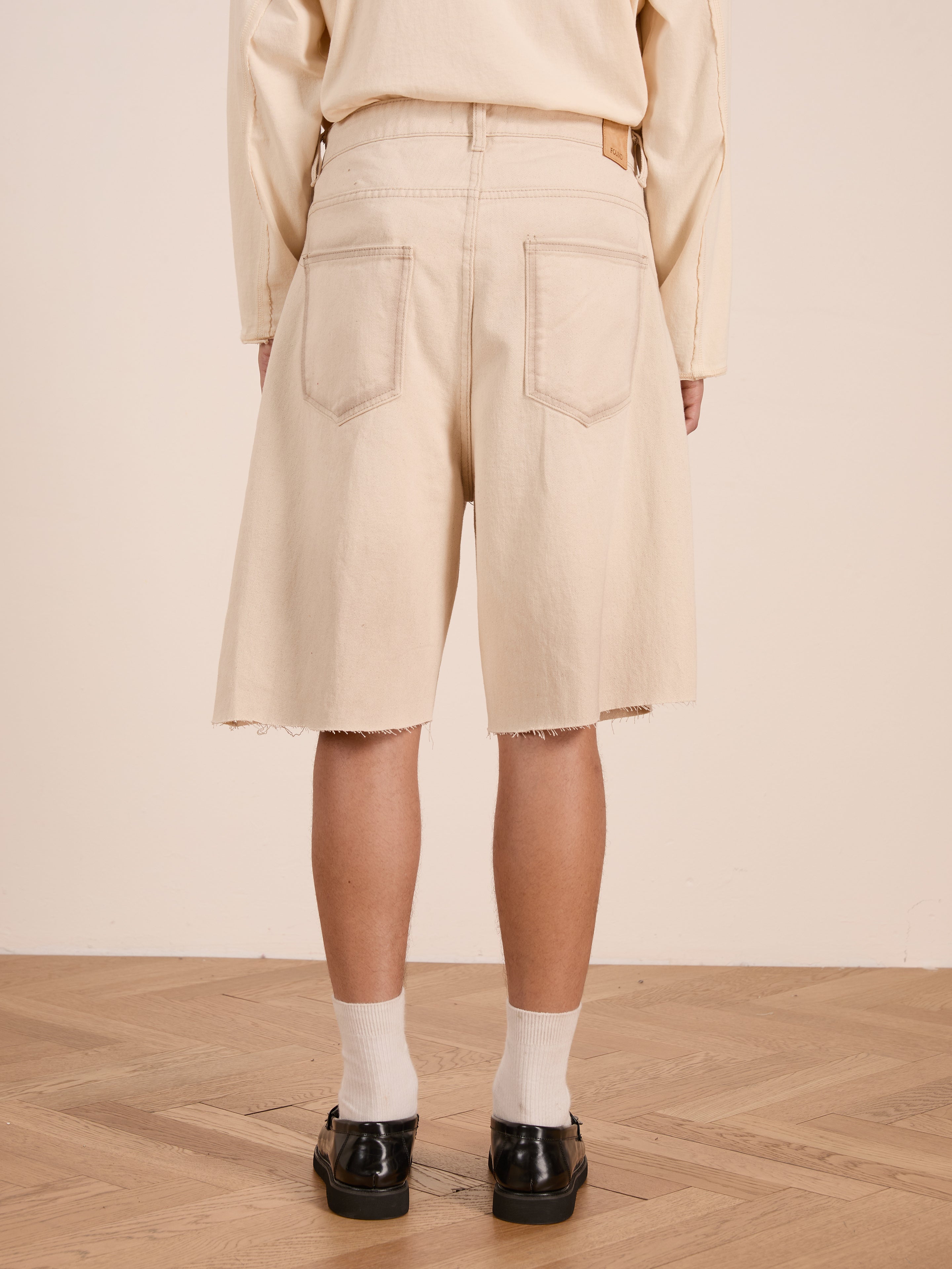 A person stands on a wooden floor, seen from the back, wearing FOUND’s Lacy Jean Jorts—relaxed fit, knee-length shorts with frayed hems—paired with white socks and black shoes for a casual unisex look.