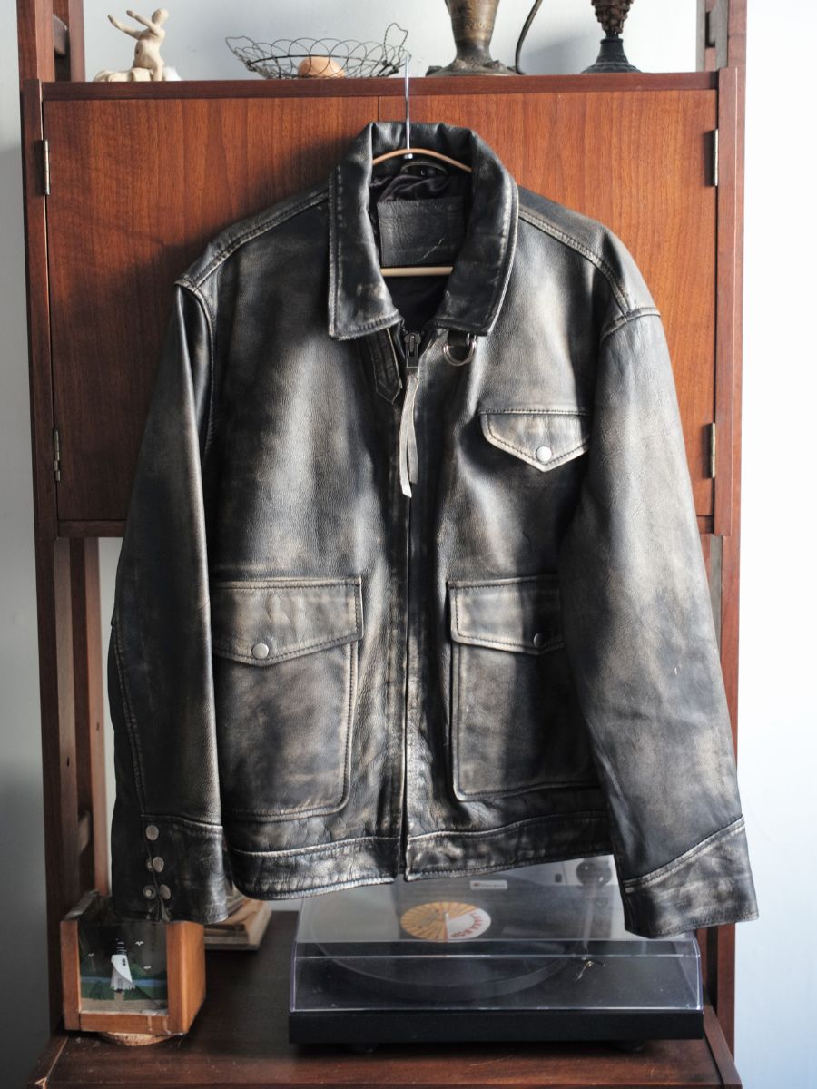 The Distressed Leather Pocket Jacket by Found, crafted from genuine cowhide, hangs on a hanger in front of a wooden cabinet with a turntable beneath.