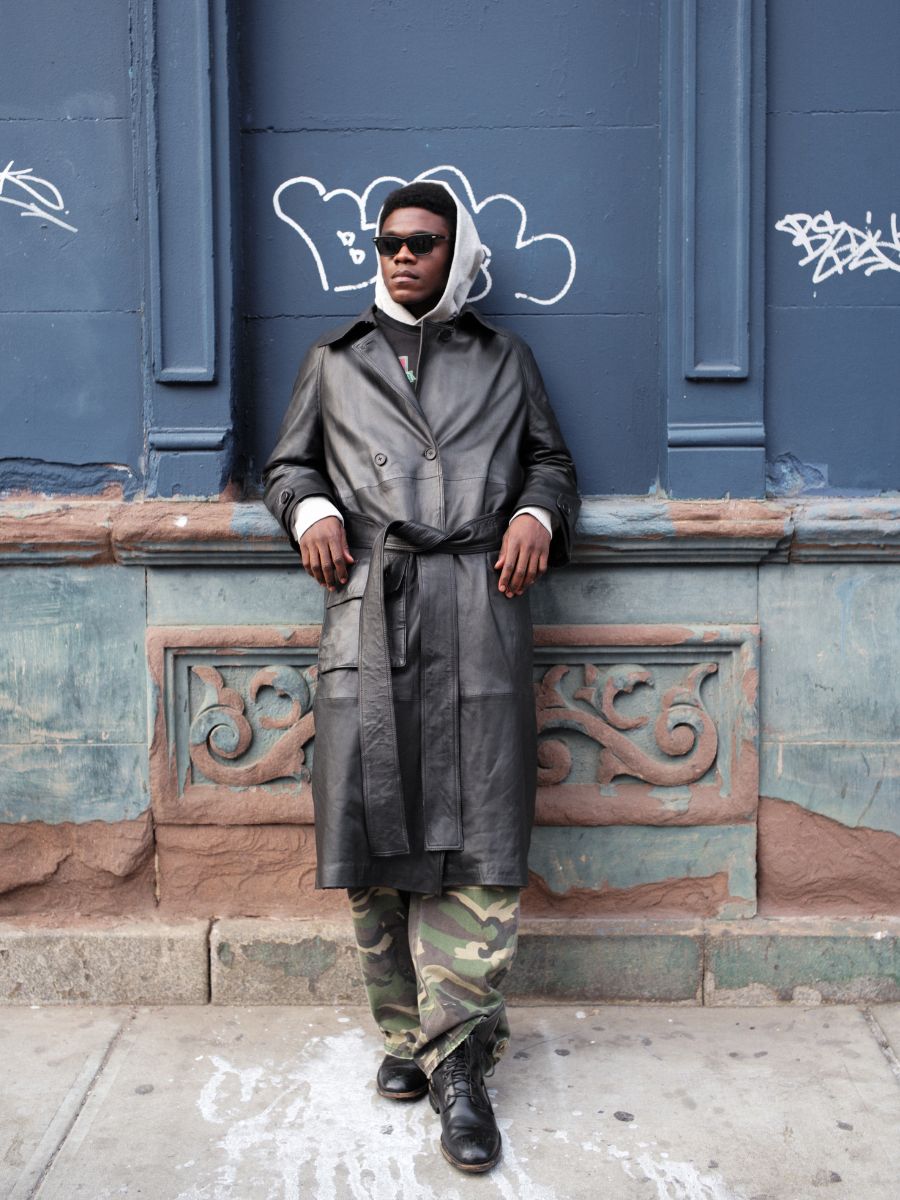 A person stands against a graffiti-covered blue wall, wearing sunglasses, a gray hoodie, camouflage pants, black boots, and the FOUND Leather Trench Coat crafted from luxurious cowhide.