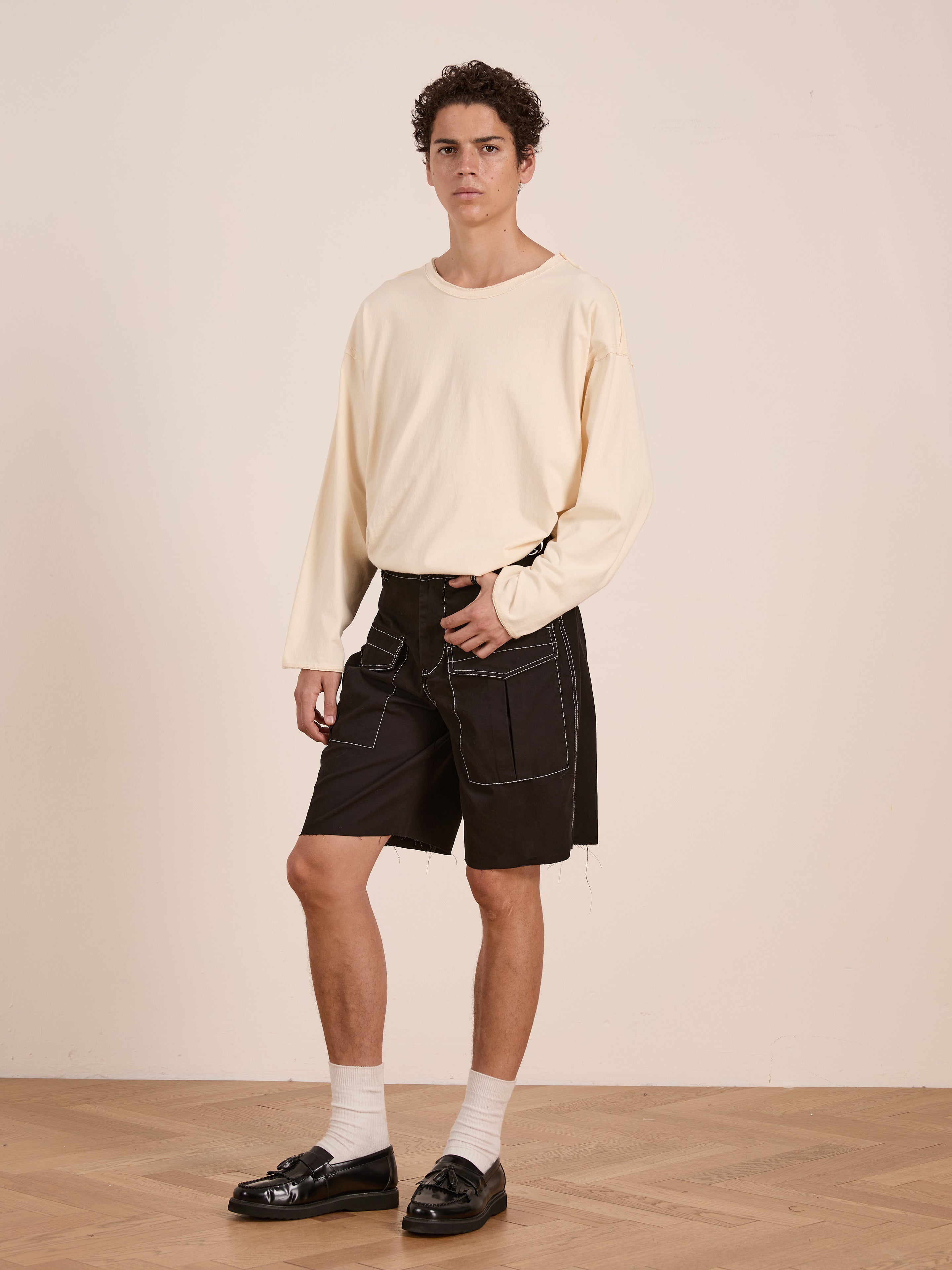 A person stands indoors in a cream long-sleeve shirt, FOUND's Lucca Black Contrast Pocket Trouser Shorts, white socks, and black loafers for a vintage-inspired look in soft cotton twill.