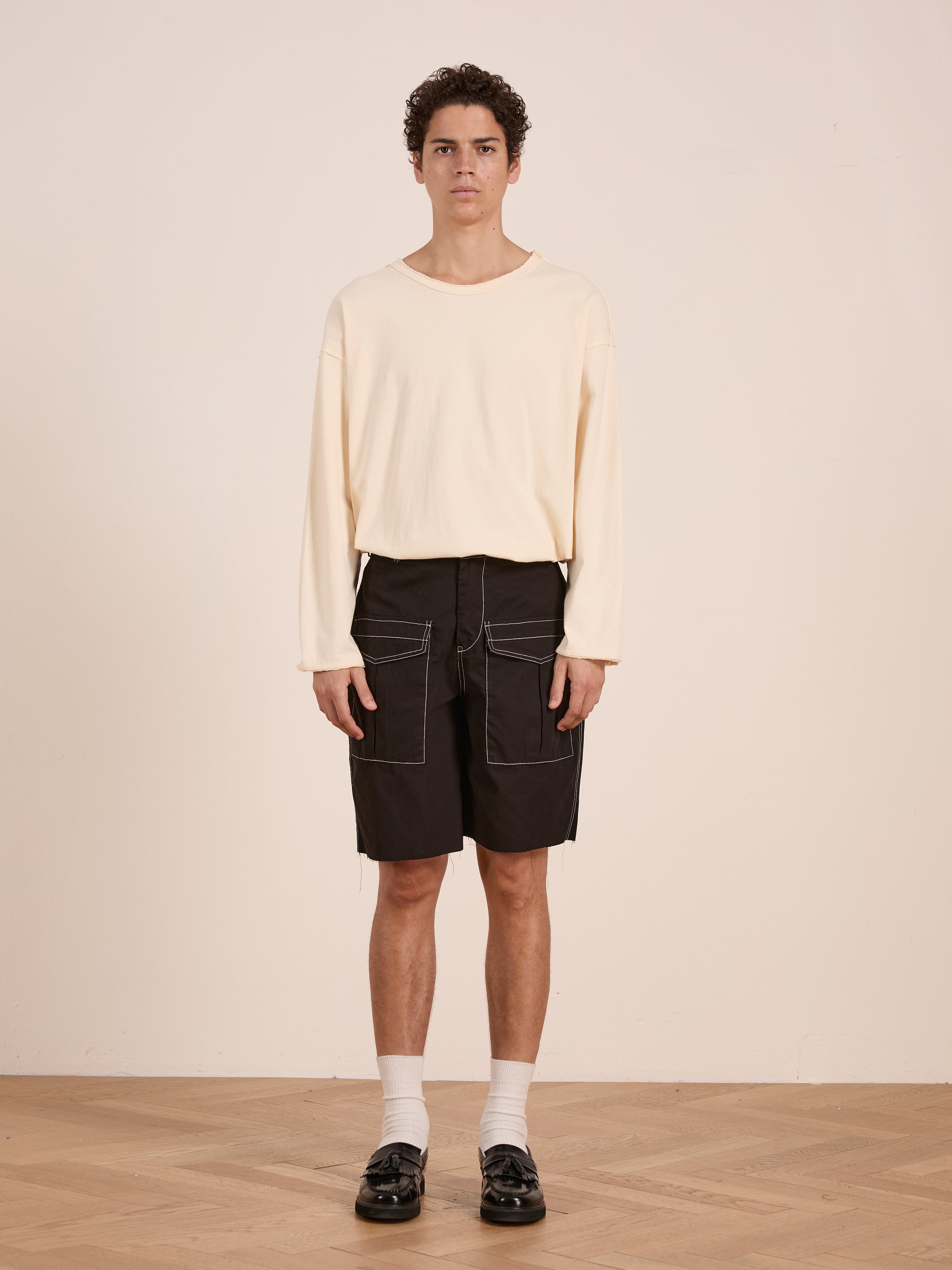 A person stands indoors against a plain wall, wearing a cream long-sleeve shirt, FOUND's Lucca Black Contrast Pocket Trouser Shorts, white socks, and black shoes for a vintage-inspired look.