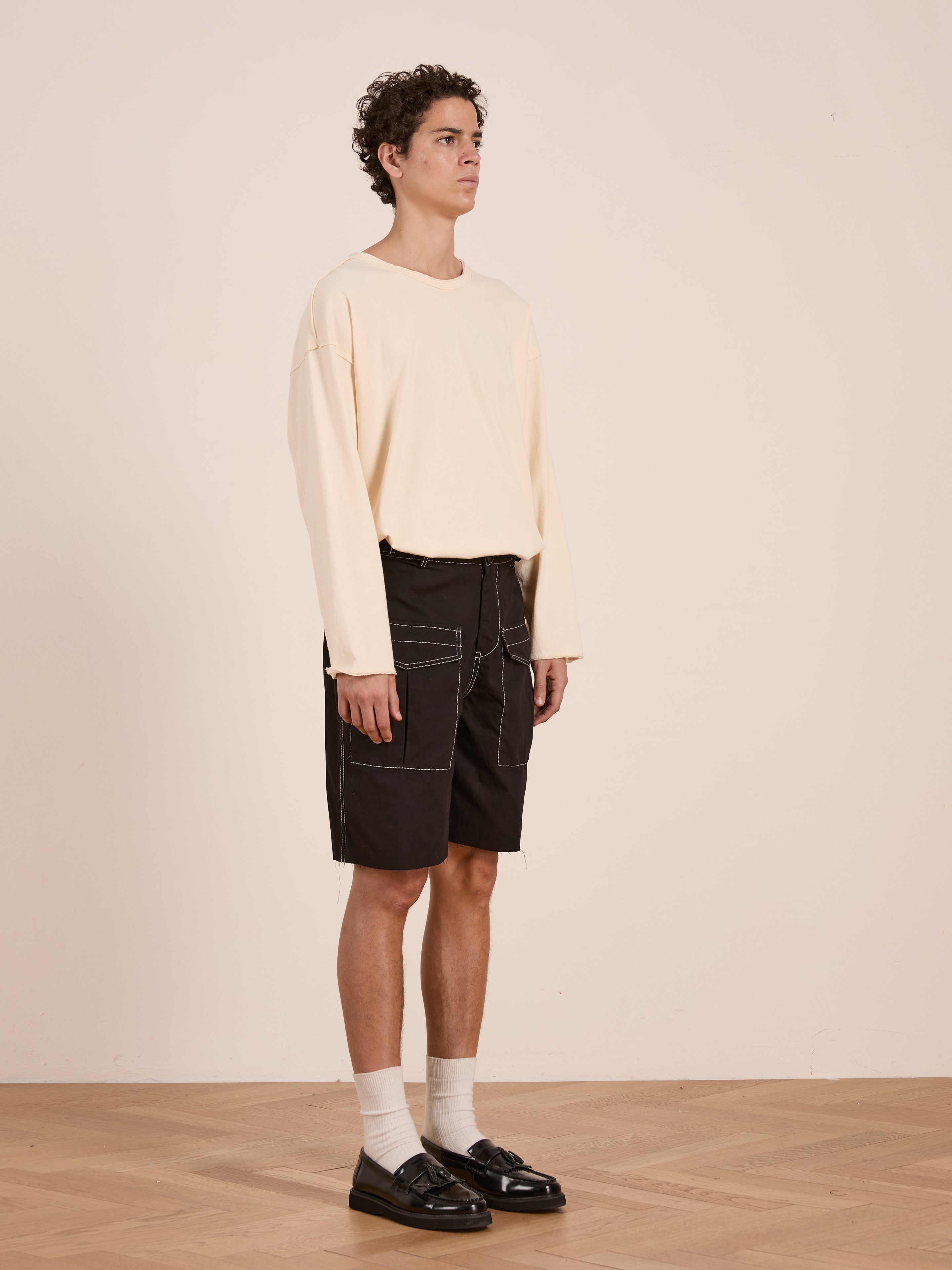 Indoors, a person wears a beige long-sleeve shirt, FOUND Lucca Black Contrast Pocket Trouser Shorts with white stitching for a vintage look, paired with white socks and black loafers on a wooden floor.