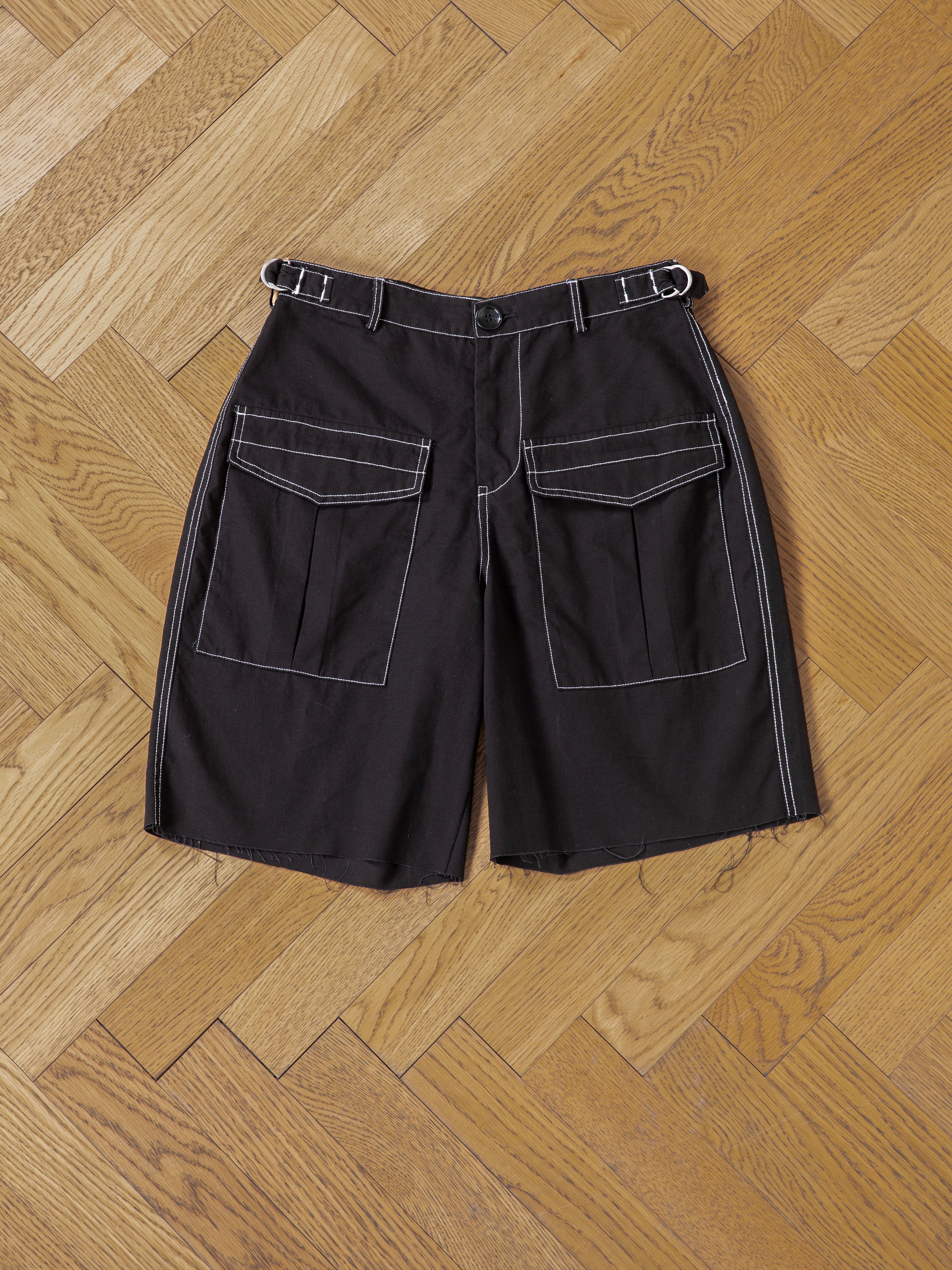 FOUND Lucca Black Contrast Pocket Trouser Shorts in cotton twill, featuring white contrast stitching, large front pockets, and adjustable waist tabs, laid flat on a wooden herringbone floor.