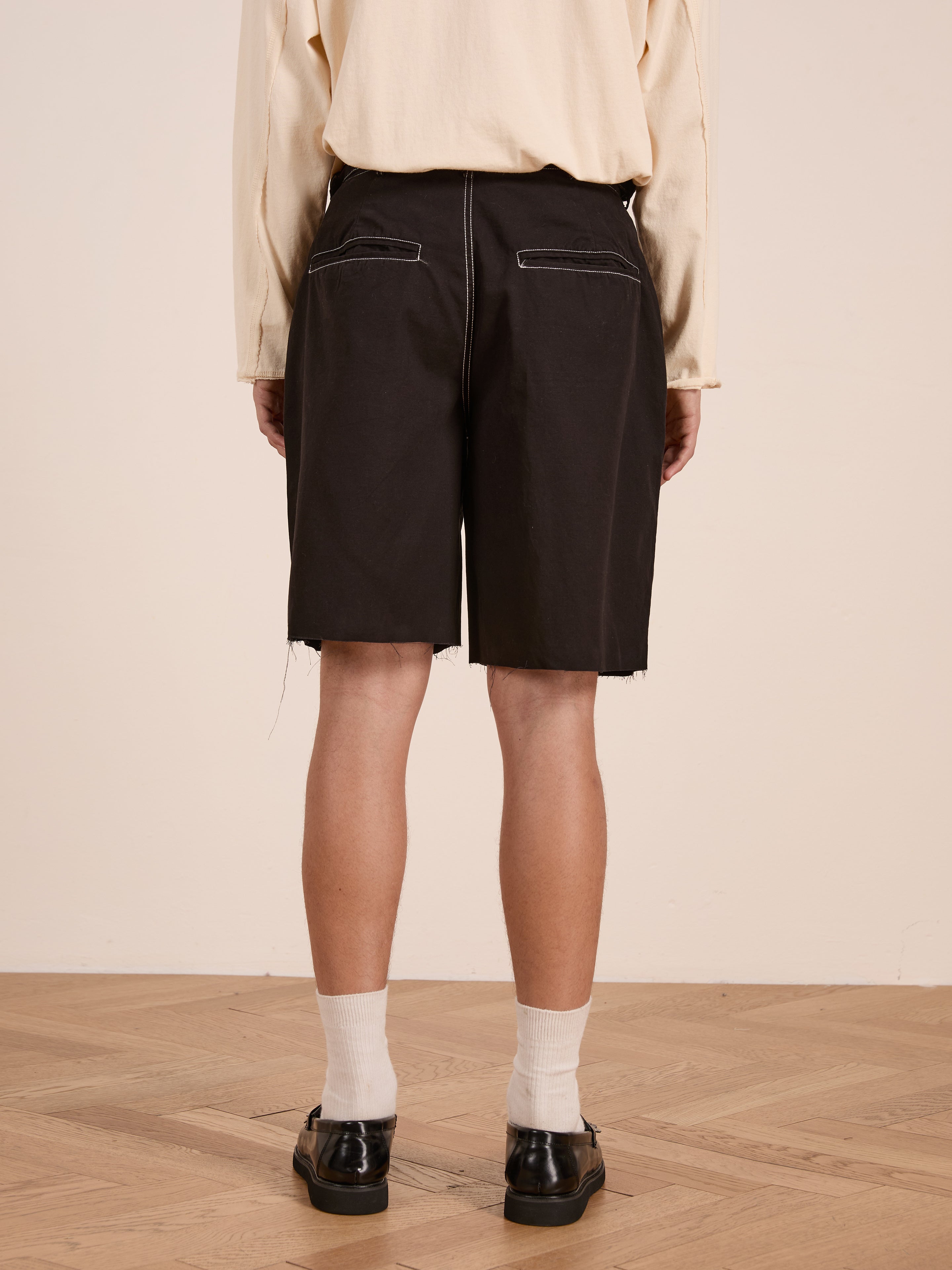 Person standing on a wooden floor, shown from the back for a vintage-inspired look, wearing FOUND's Lucca Black Contrast Pocket Trouser Shorts in cotton twill with white stitching, beige long-sleeve shirt, white socks, and black shoes.