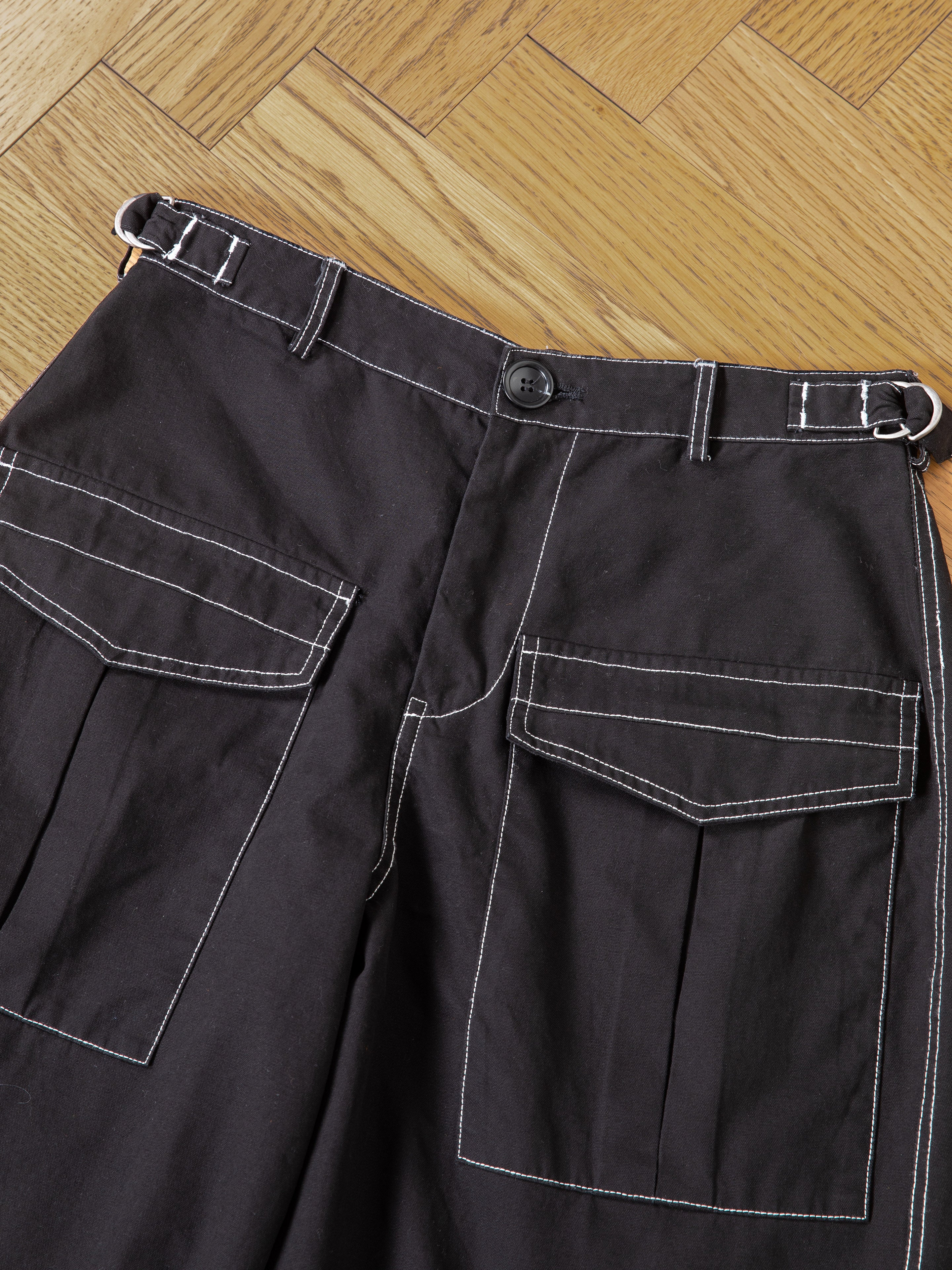 Close-up of FOUND Lucca Black Contrast Pocket Trouser Shorts, featuring large flap pockets and white contrast stitching, laid flat on a herringbone wooden floor for a vintage-inspired look.