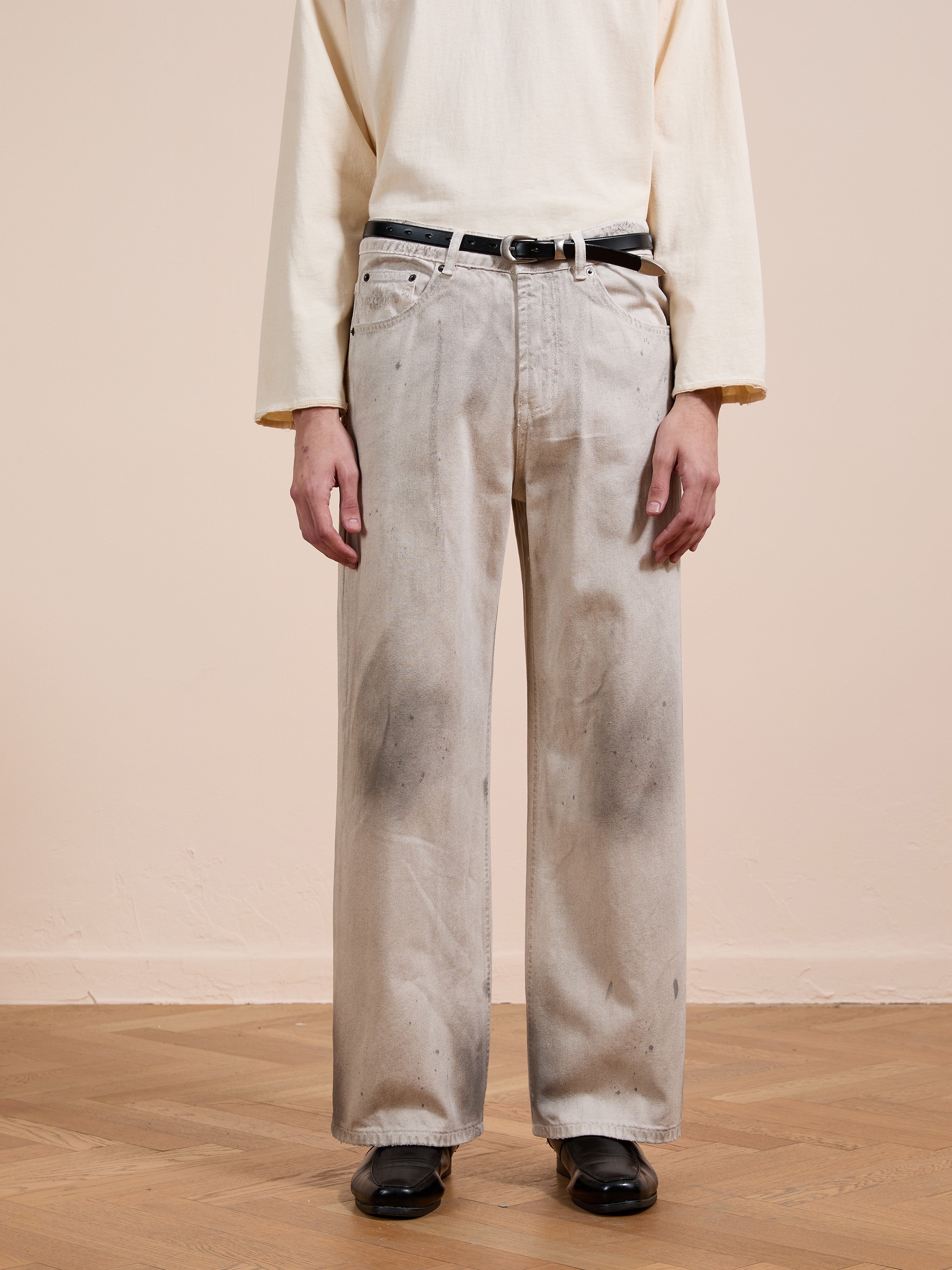 A person wears FOUND's Lacy Baggy Jeans—unisex, relaxed fit, wide-leg, distressed concrete wash with dark stains—paired with a beige long-sleeve shirt, black belt and shoes, standing on a wooden floor against a neutral wall.