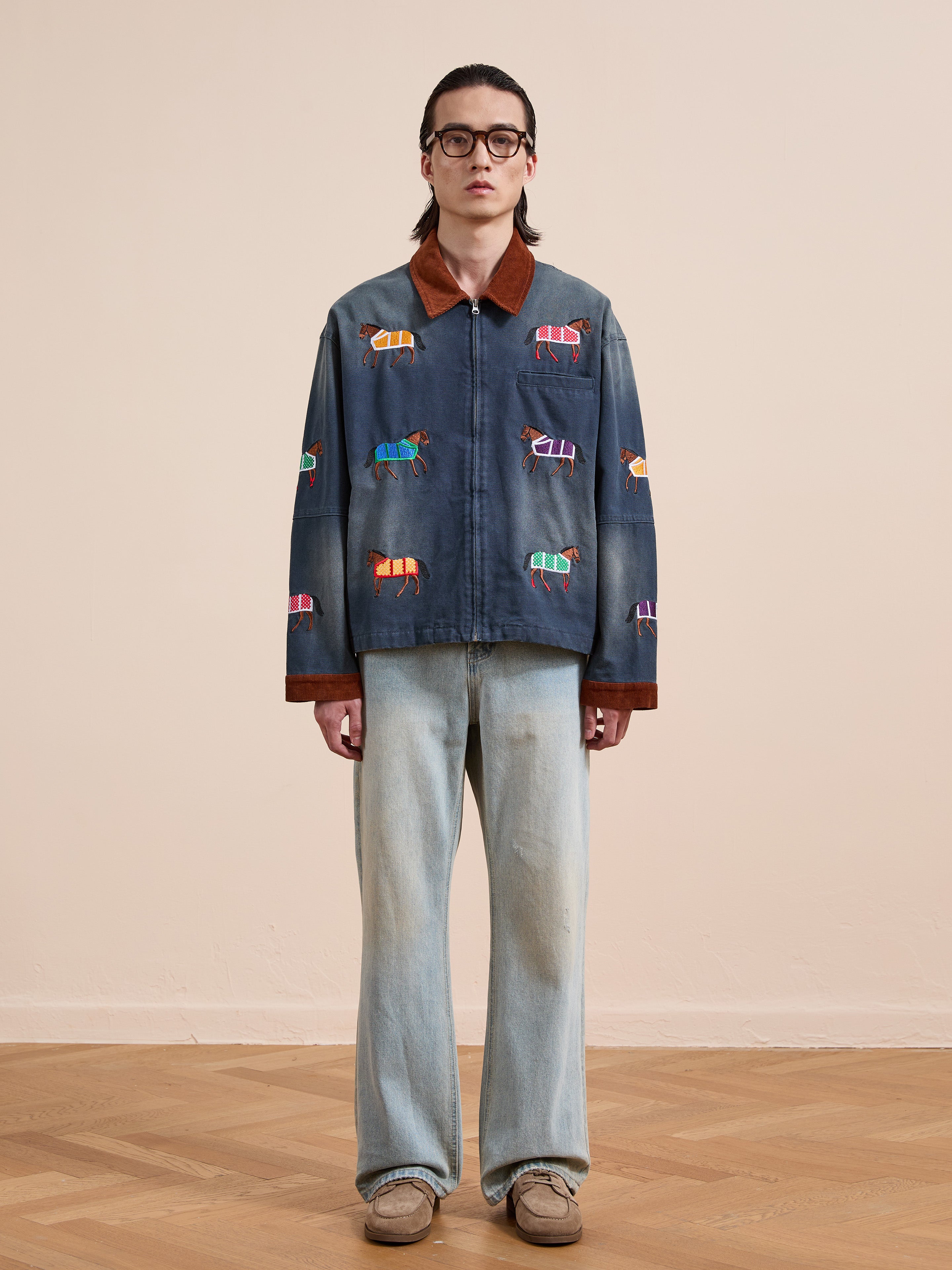 Indoors, a person wears glasses, light blue jeans, beige shoes, and the Found Lariat Horse Embroidered Work Jacket, a unisex navy design with colorful horse embroidery, standing against a plain background.