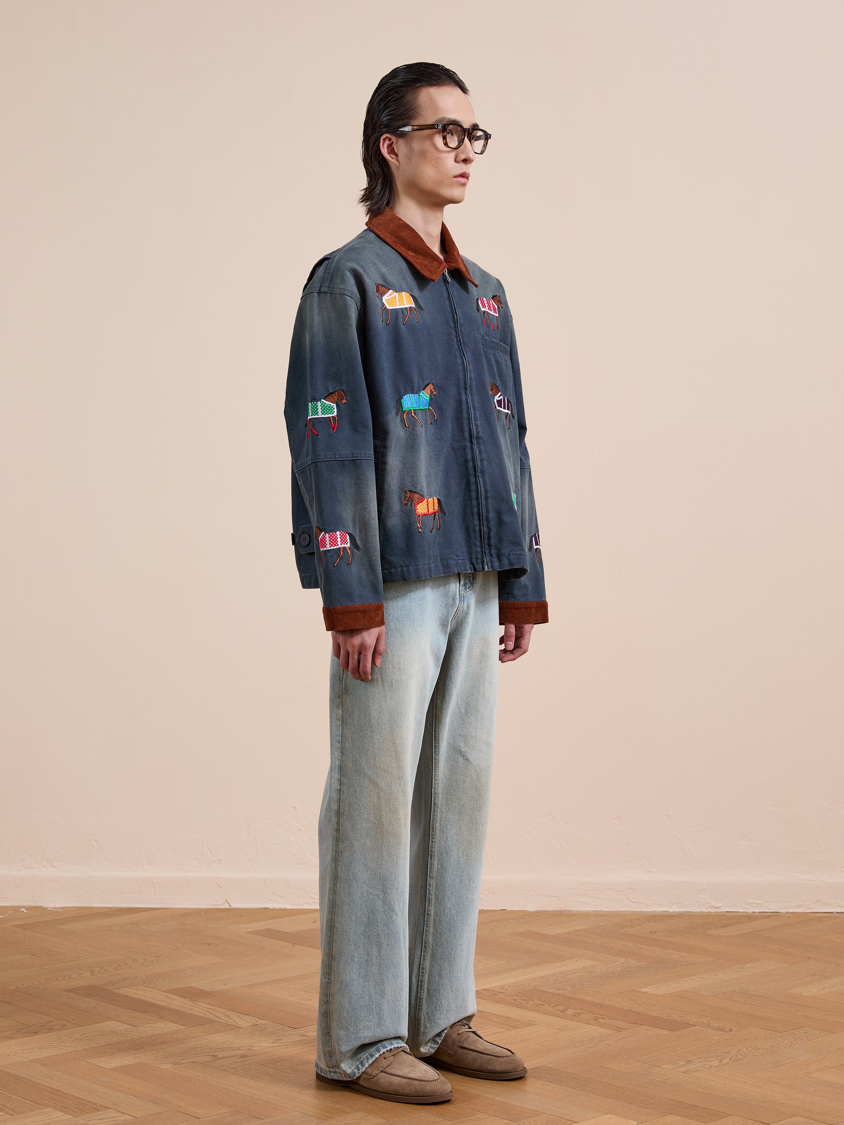 A person stands indoors on a wooden floor wearing glasses, light jeans, brown shoes, and the Found Lariat Horse Embroidered Work Jacket with animal motifs.