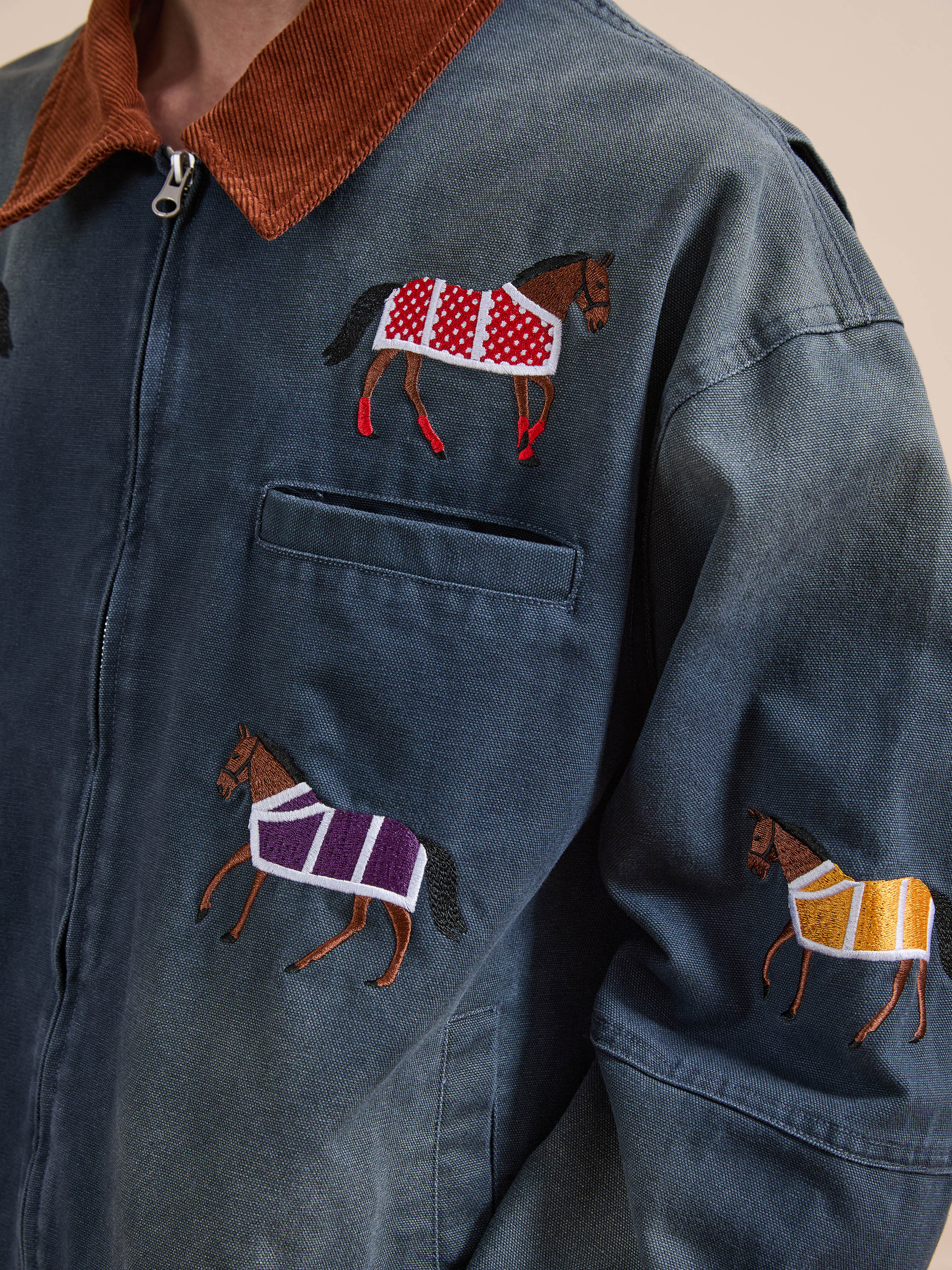 A close-up of Found's Lariat Horse Embroidered Work Jacket—this denim jacket features intricate horse embroidery in red, purple, and yellow blankets and is finished with a brown corduroy collar.